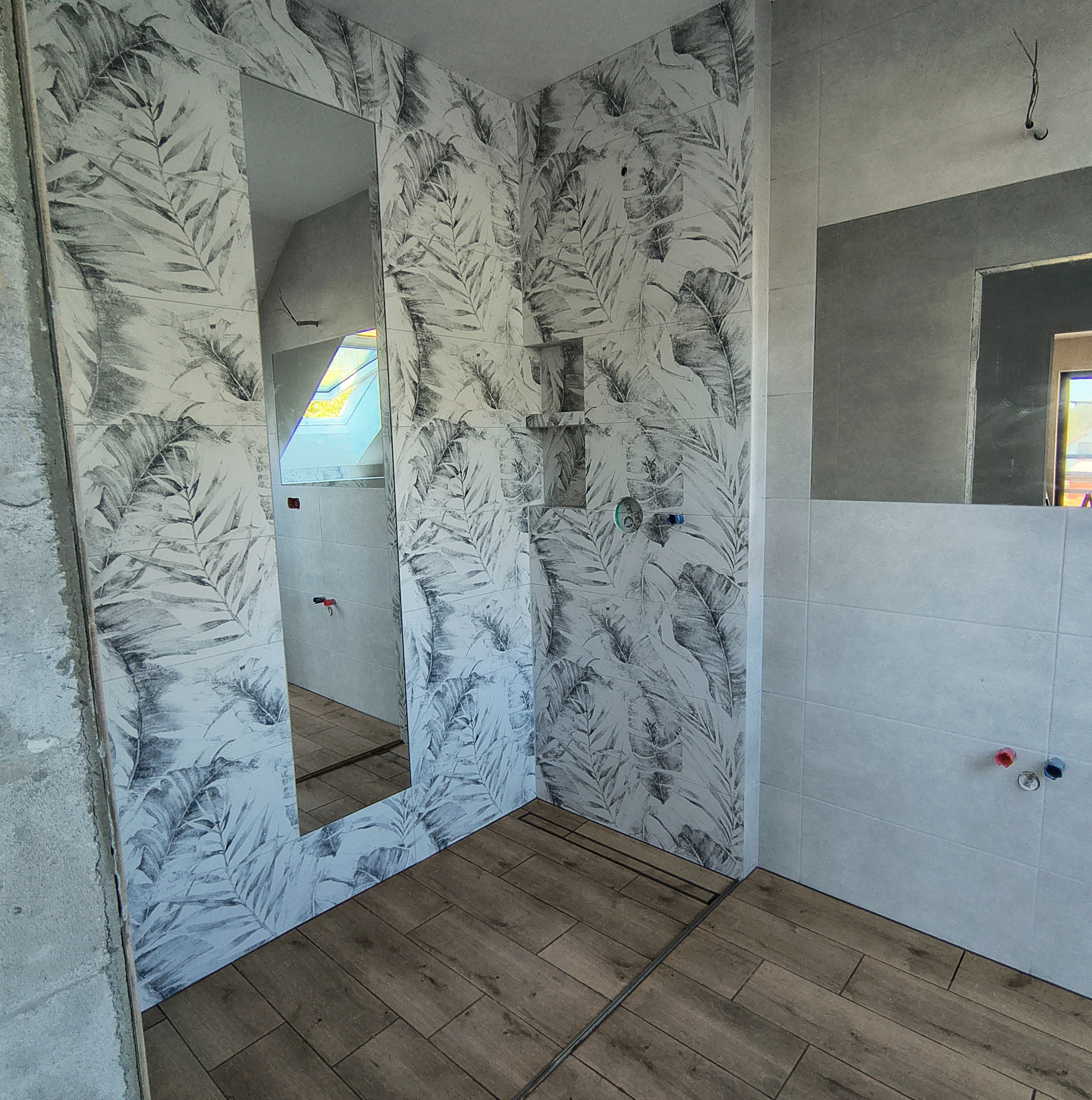 Wykończona łazienka z drewnopodobną podłogą, ściany w biało-szare wzory liści. Widoczne lustra i elementy instalacji. Częściowo wykończone obrzeża.