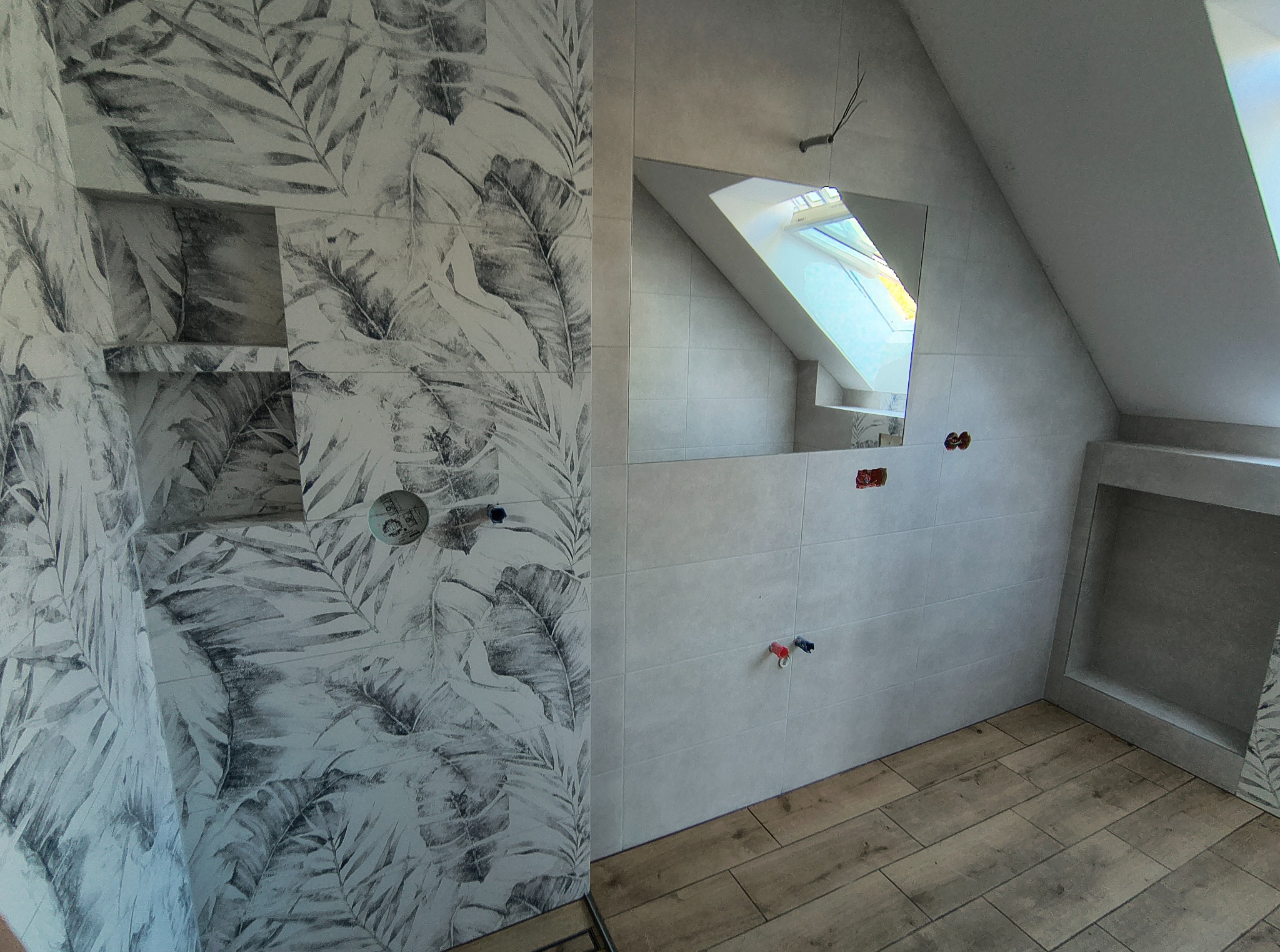 Wykończona łazienka na poddaszu z płytkami w liście, drewnopodobną podłogą i lustrem odbijającym okno dachowe. Widoczne instalacje wodne.