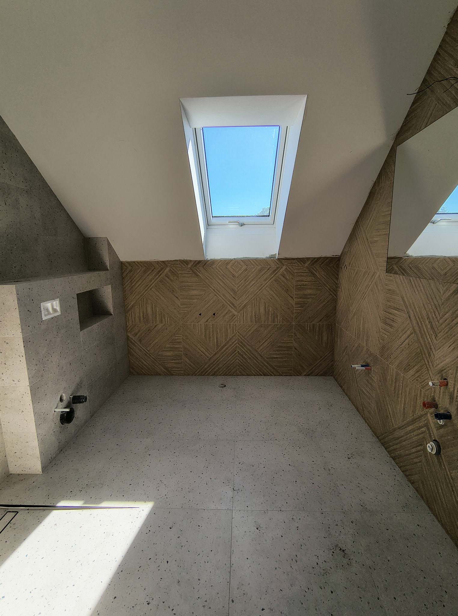 Nowoczesna łazienka na poddaszu z oknem dachowym i geometrycznymi płytkami ściennymi. Widoczne przygotowane instalacje wodne i kanalizacyjne, surowy beton i szara podłoga.