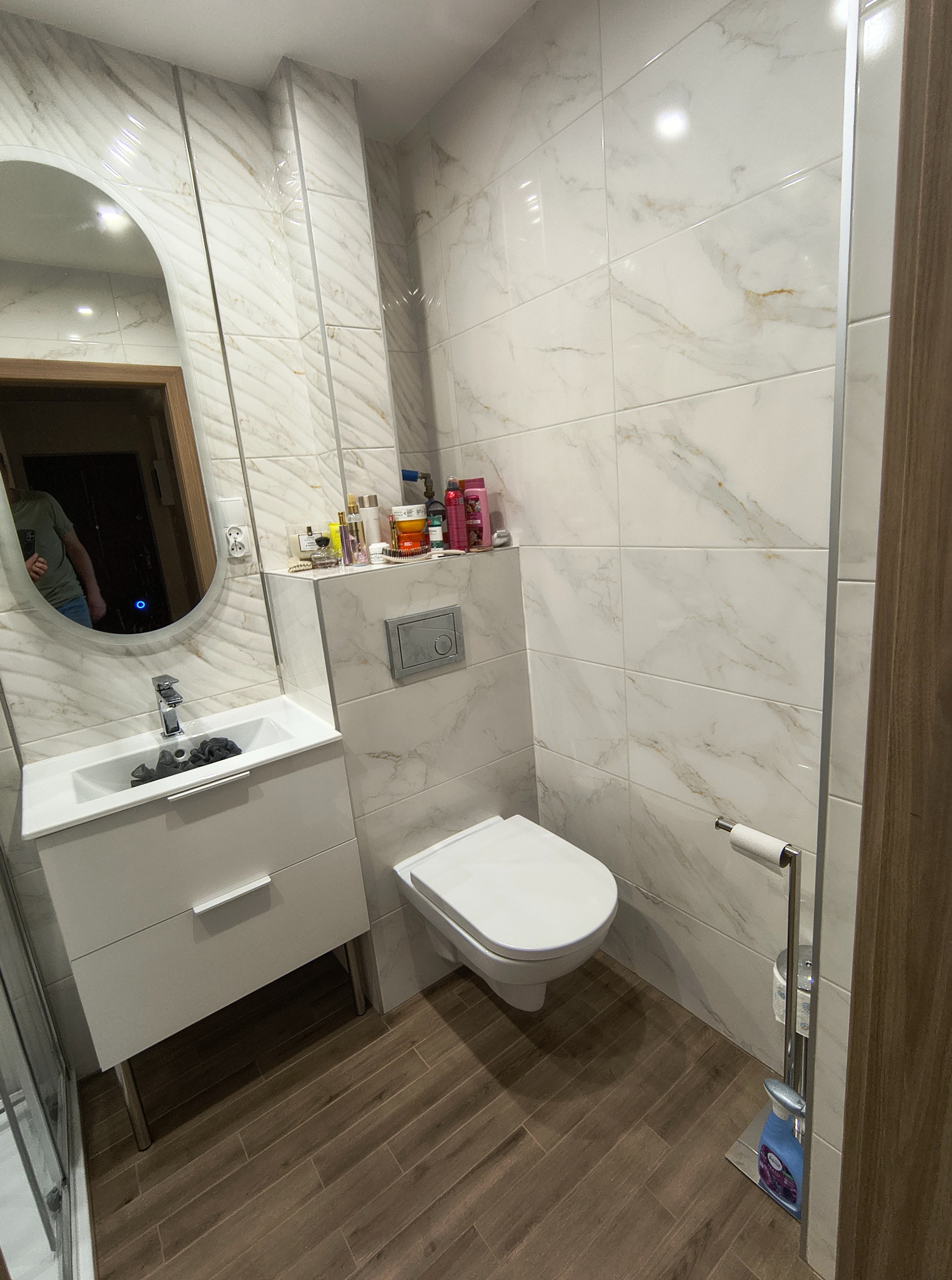 Jasna łazienka po renowacji: białe marmurowe płytki, drewnopodobna podłoga, owalne lustro, biała szafka z umywalką, toaleta i akcesoria.