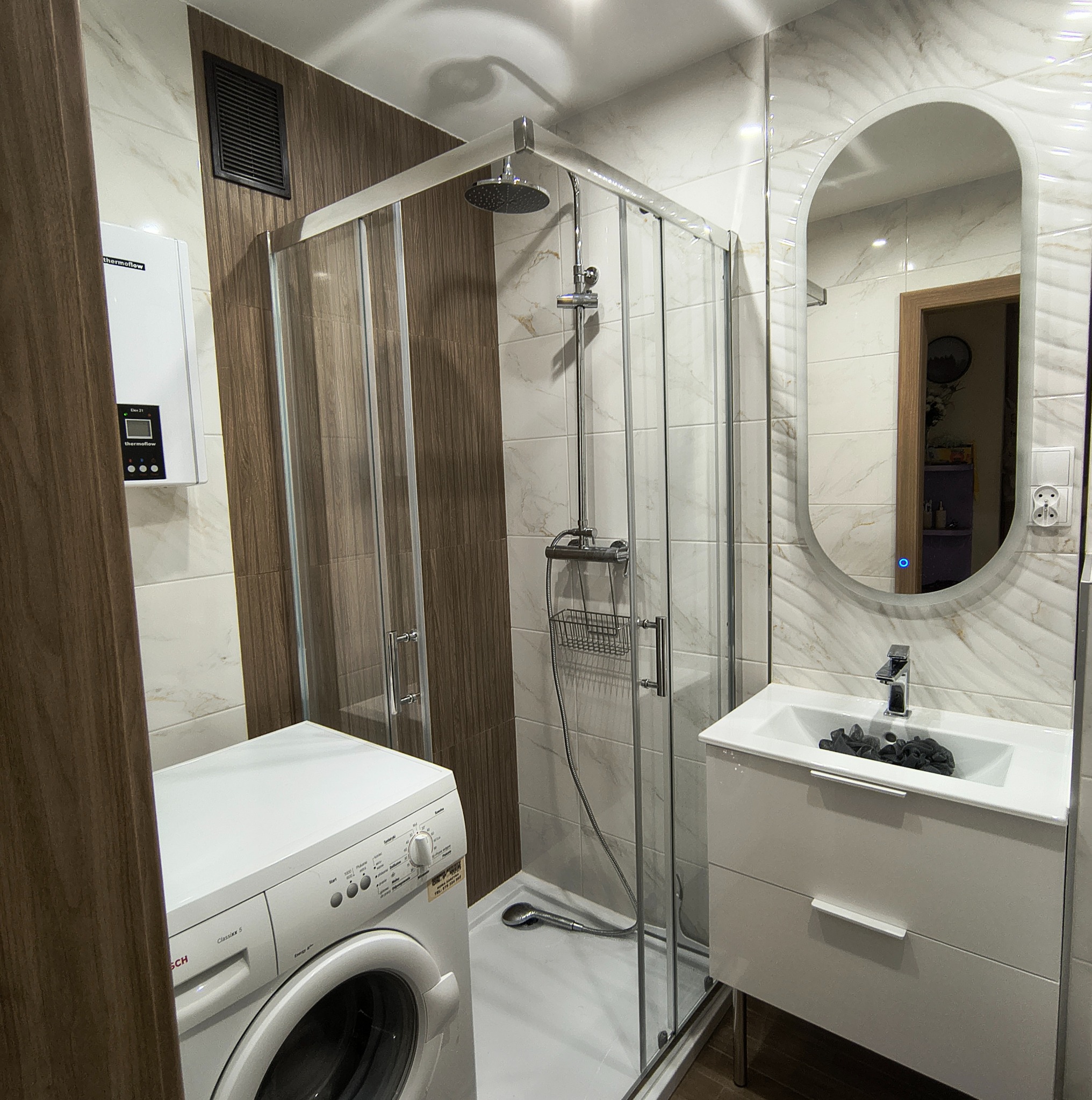 Nowoczesna łazienka z kabiną prysznicową, białą umywalką z szafką i pralką Bosch. Marmurowe płytki i podświetlane lustro owalne dopełniają wystrój.