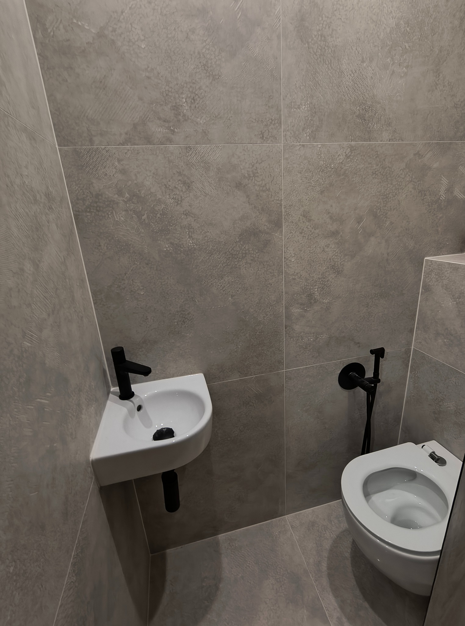 Nowoczesna, szara łazienka z wiszącą toaletą i narożną umywalką. Czarna armatura wyróżnia się na tle ścian z efektem betonu.