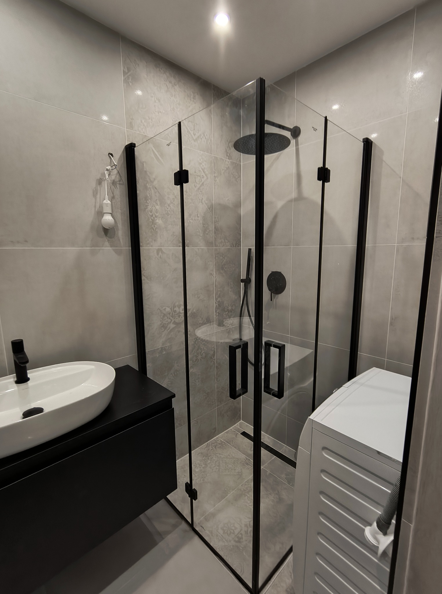 Nowoczesna łazienka z kabiną prysznicową typu walk-in z czarnymi profilami, umywalką nablatową i pralką. Szare płytki na ścianach i podłodze.