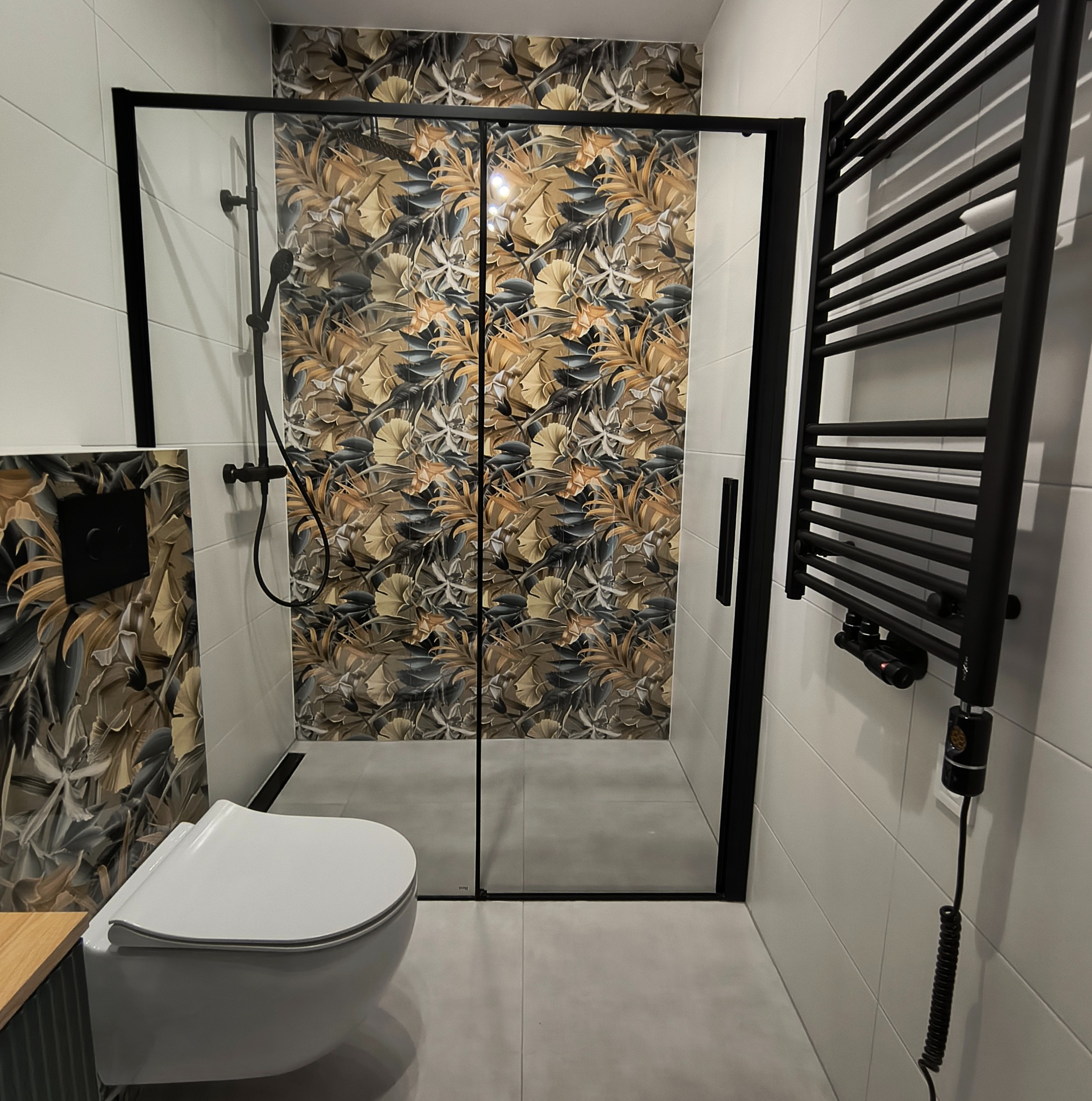 Wykończona łazienka z kabiną prysznicową typu walk-in, toaletą podwieszaną i grzejnikiem drabinkowym w czarnych ramach. Ściana w kabinie z motywem liści. Nowoczesny design.