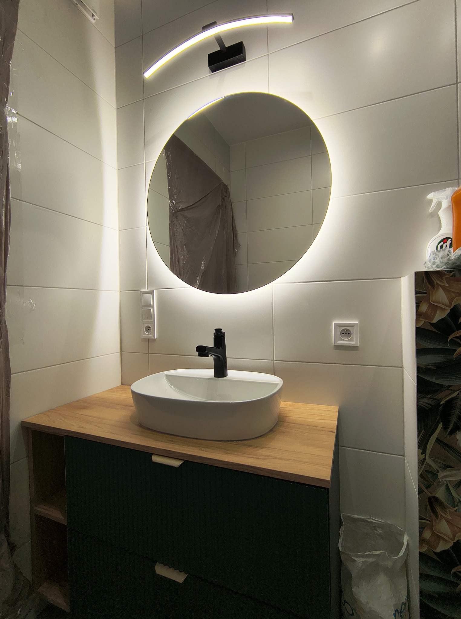 Nowoczesna łazienka z okrągłym lustrem LED, białą ceramiką i drewnianym blatem. Minimalistyczny design, jasne kafelki, czarna armatura. Częściowo widoczna folia ochronna.