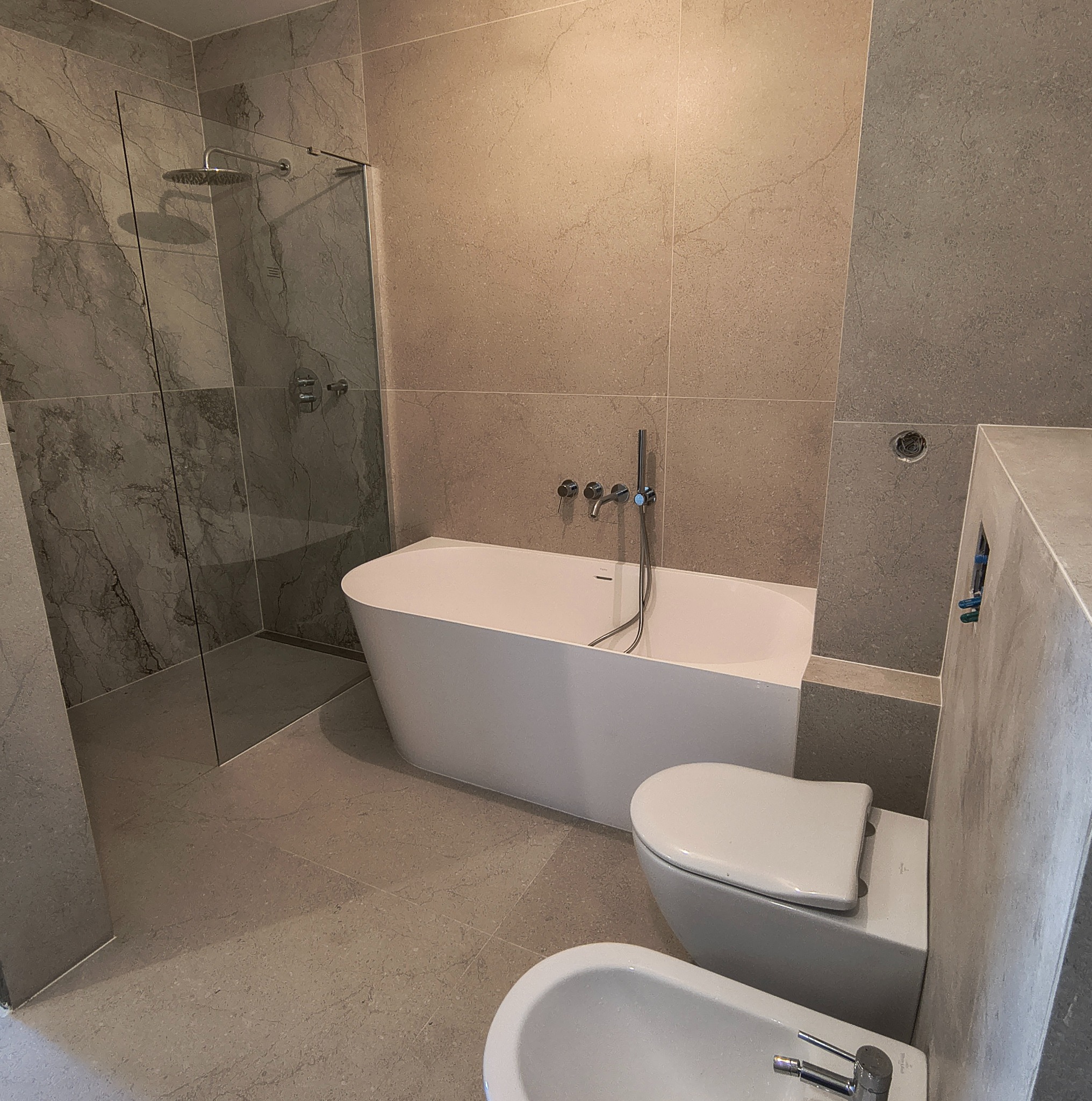 Nowoczesna łazienka z kabiną prysznicową typu walk-in, wolnostojącą białą wanną, toaletą i bidetem. Płytki w odcieniach szarości imitujące kamień. Minimalistyczne baterie.