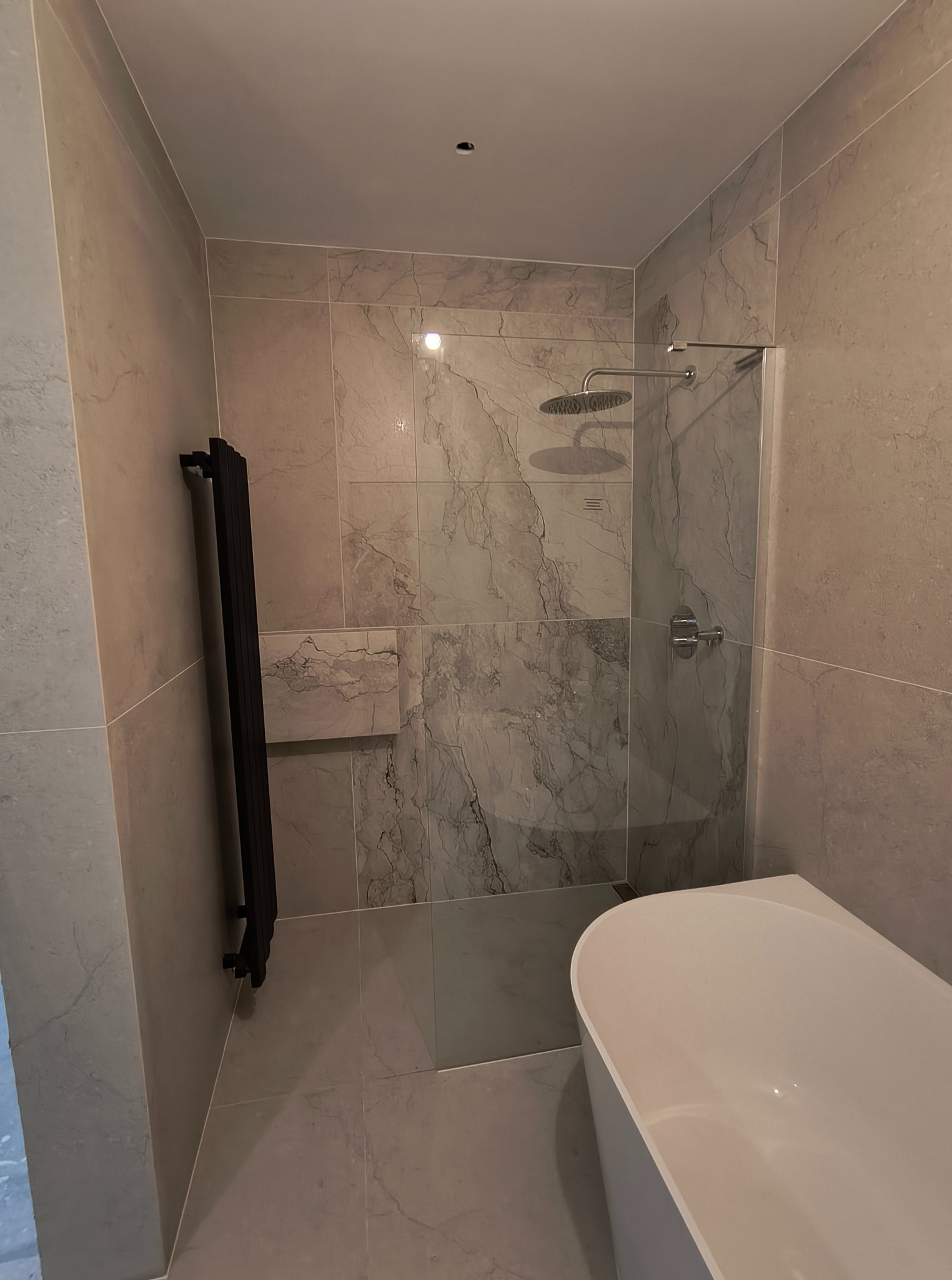 Nowoczesna łazienka z marmurowymi płytkami, czarnym grzejnikiem drabinkowym i białą wanną. Widoczna szklana kabina prysznicowa z deszczownicą.