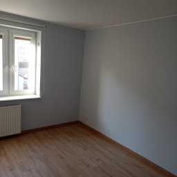 Tomasz Kruszka - Pomalowane na szaro wnętrze pokoju z drewnianą podłogą, widoczne okno z grzejnikiem i nowoczesna lampa sufitowa. Ściany gładkie, gotowe do aranżacji.