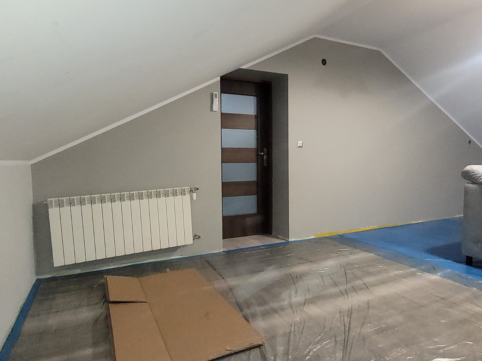 Pokój na poddaszu po odświeżeniu: szare ściany, biały grzejnik, drewniane drzwi z przeszkleniami. Podłoga zabezpieczona folią malarską.