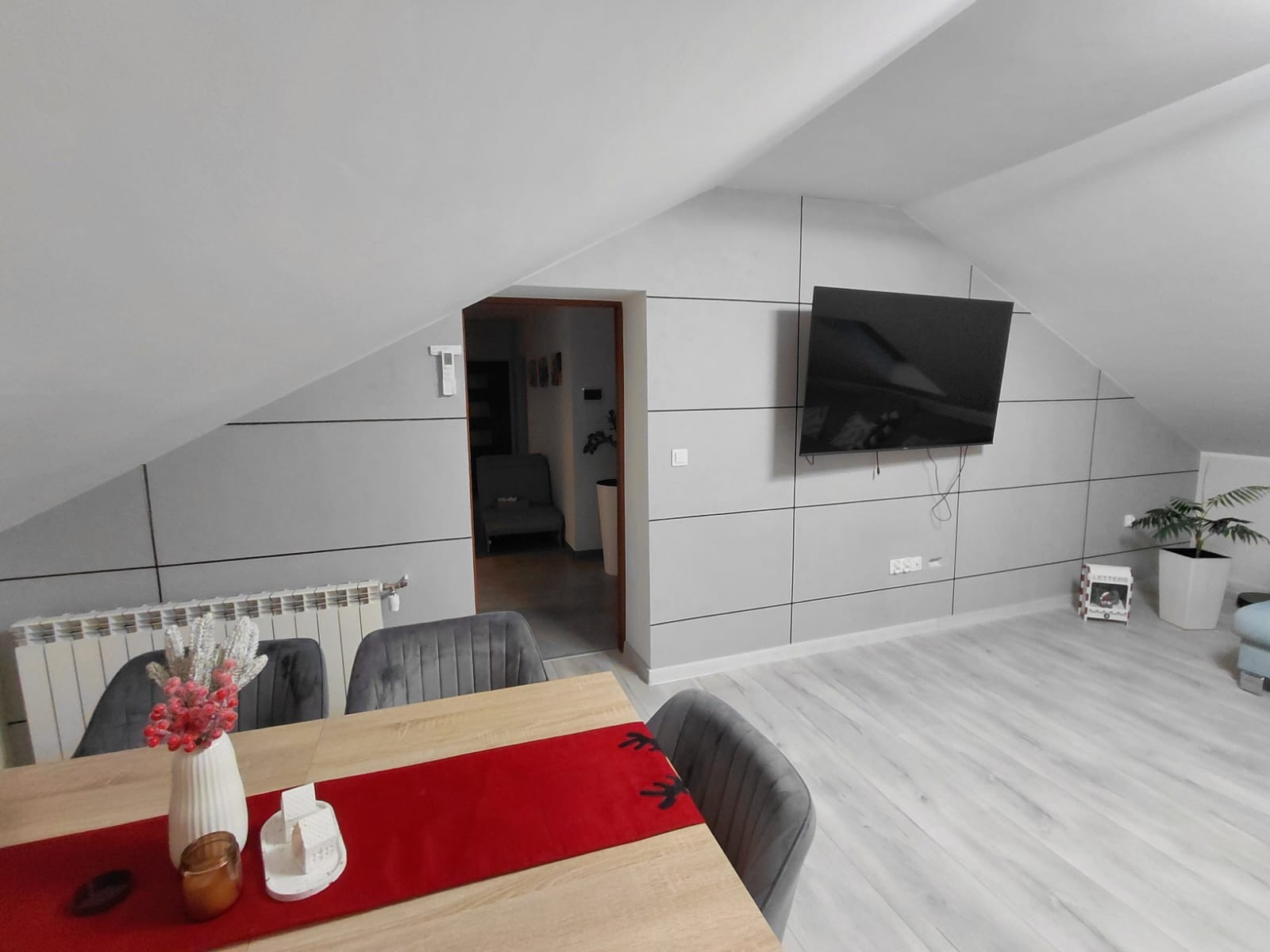 Jasne wnętrze poddasza po remoncie: szare ściany z panelami, telewizor, stół z dekoracjami, widok na salon. Nowoczesny design i wykończenie.