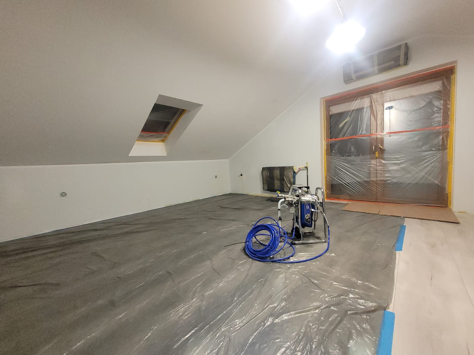 Przygotowane poddasze do prac wykończeniowych w Tczewie. Zabezpieczona folią podłoga, okno i drzwi. Widoczny agregat malarski.
