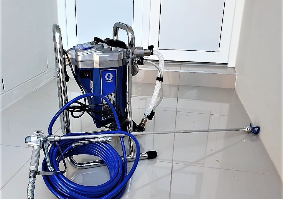 Agregat malarski Graco na podłodze z płytek, gotowy do użycia. Widoczny pistolet z niebieskim wężem i zbiornik na farbę. Sprzęt profesjonalny do malowania natryskowego.