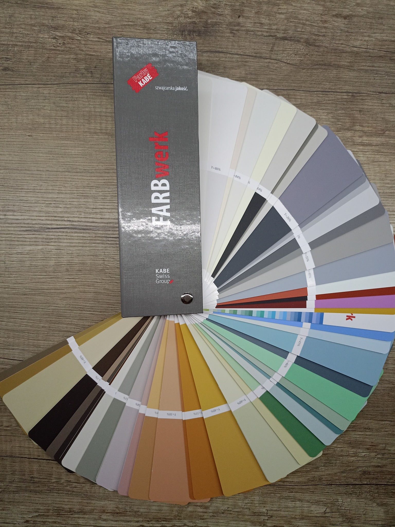 Paleta kolorów Farby KABE na drewnianym tle. Wachlarz odcieni bieli, szarości, beżu, brązu, zieleni i błękitu. Katalog z próbkami kolorów do wyboru przy malowaniu.