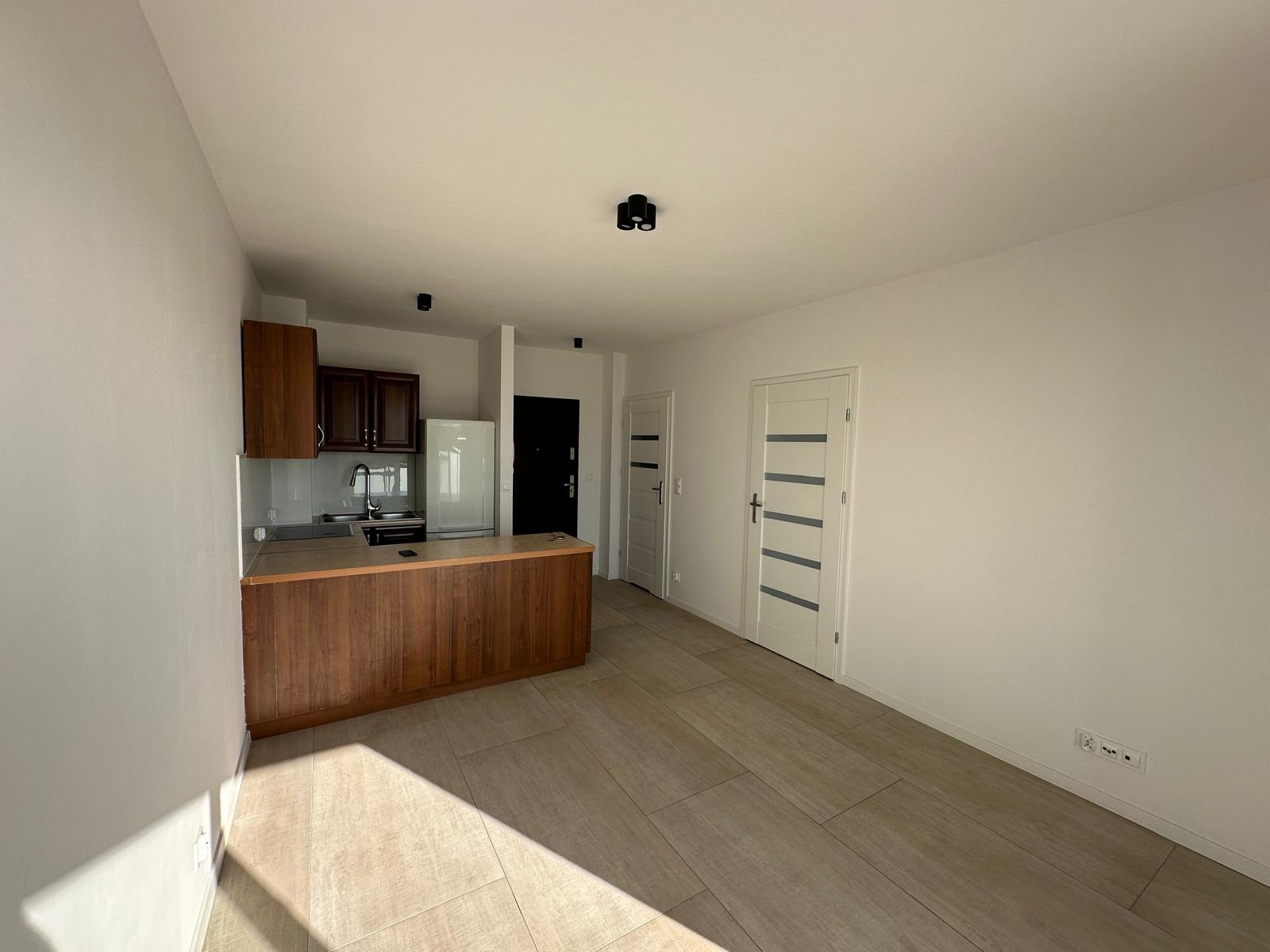 Jasne wnętrze po remoncie: białe ściany, drewniana podłoga i zabudowa kuchenna. Widoczne gładkie powierzchnie ścian i sufitu, nowe drzwi wewnętrzne.