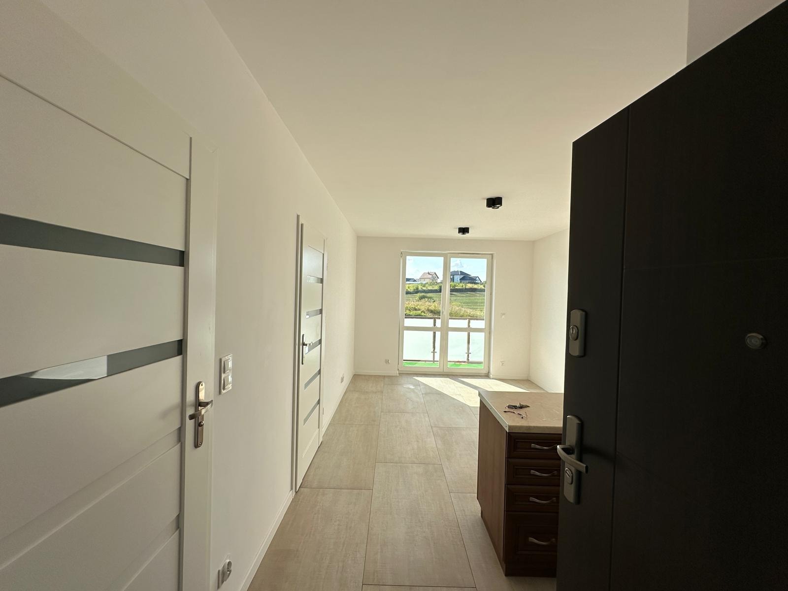 Jasne wnętrze po remoncie: białe ściany, drzwi z przeszkleniami, beżowa podłoga, okno z widokiem na zieleń. Ciemne drzwi wejściowe po prawej stronie kadru.