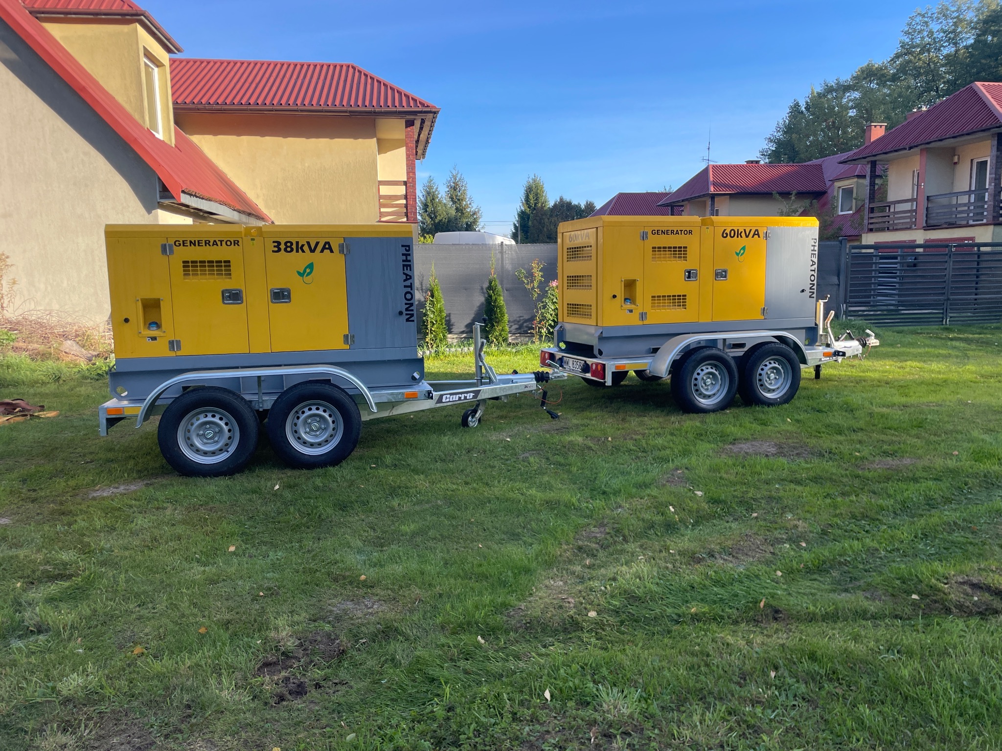 Dwa generatory mobilne (38kVA i 60kVA) na przyczepach, marki Pheatonn, stoją na trawie przed budynkami z czerwoną dachówką w słoneczny dzień.