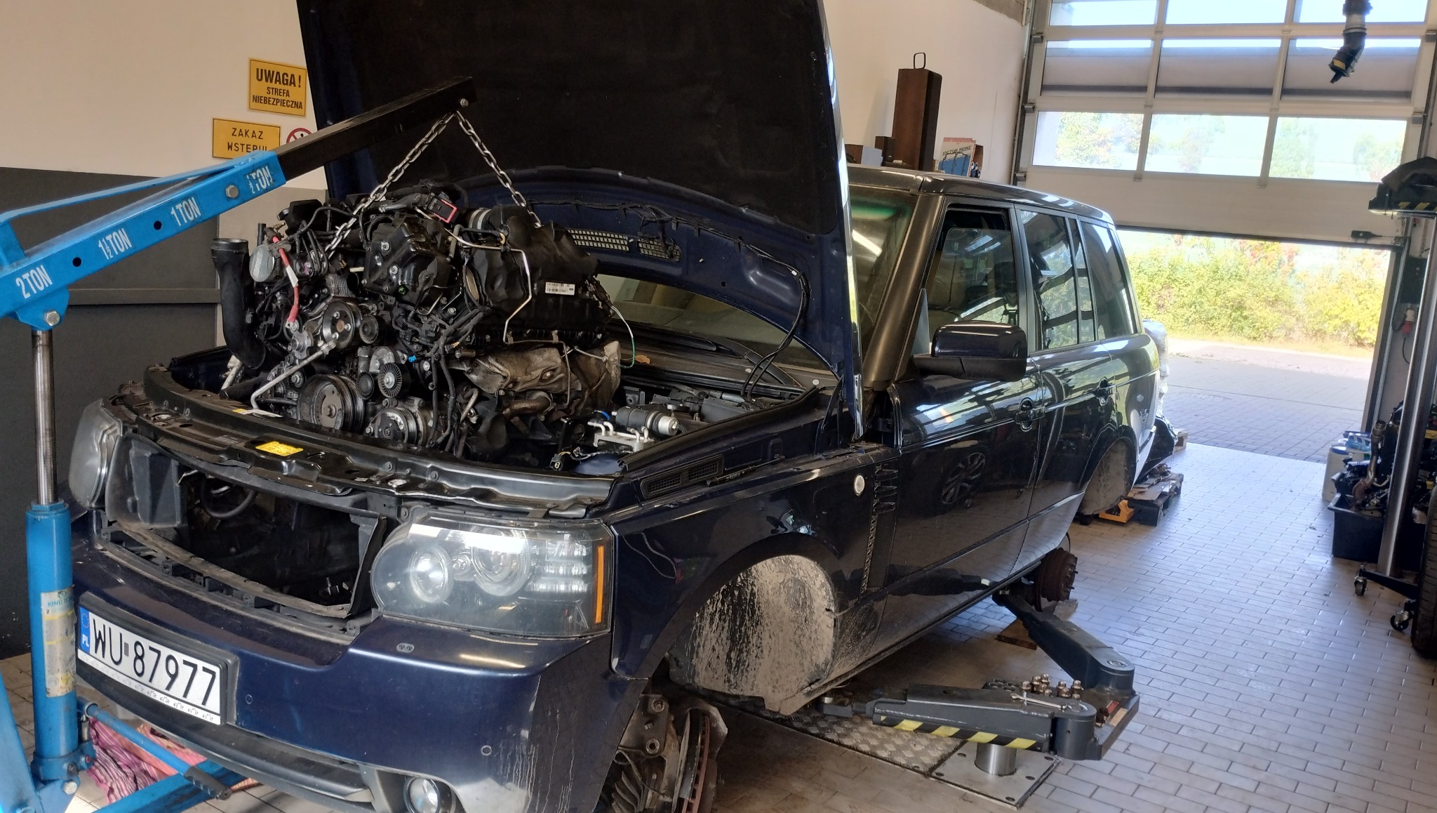 Niebieski Range Rover na podnośniku w warsztacie, z otwartą maską i wyjętym silnikiem, podwieszonym na suwnicy. Widoczne narzędzia i tablice ostrzegawcze.