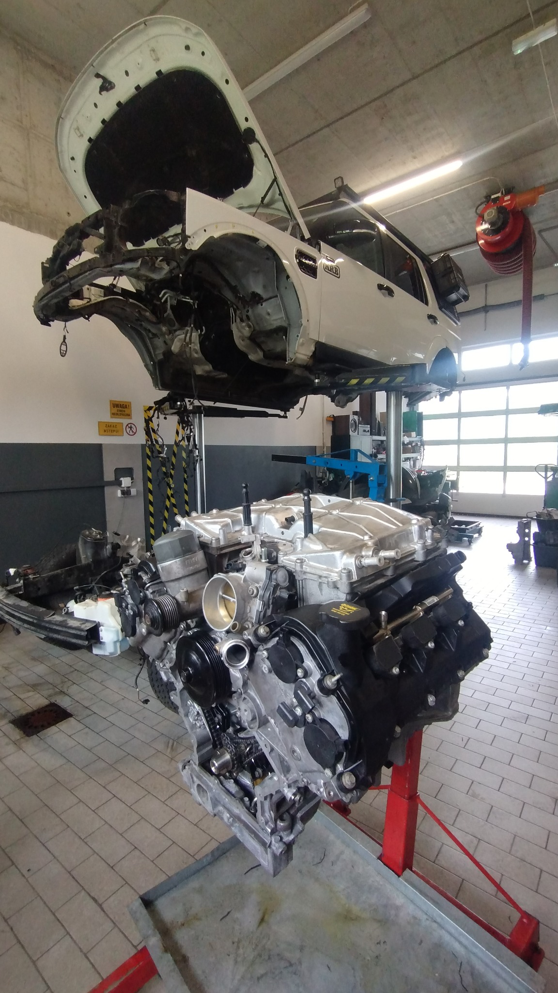 Warsztat samochodowy: biały SUV na podnośniku, zdemontowany silnik na stojaku. Widoczne detale silnika, elementy karoserii. Prace mechaniczne w toku.