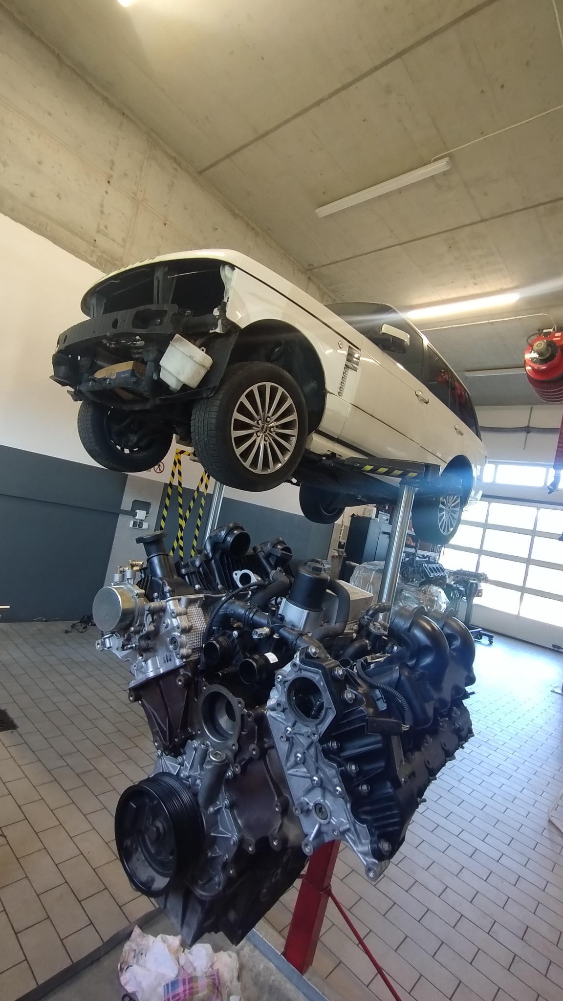 Biały samochód na podnośniku warsztatowym, obok silnik na wózku. Widoczne elementy wnętrza warsztatu, takie jak podłoga z płytek i brama garażowa.