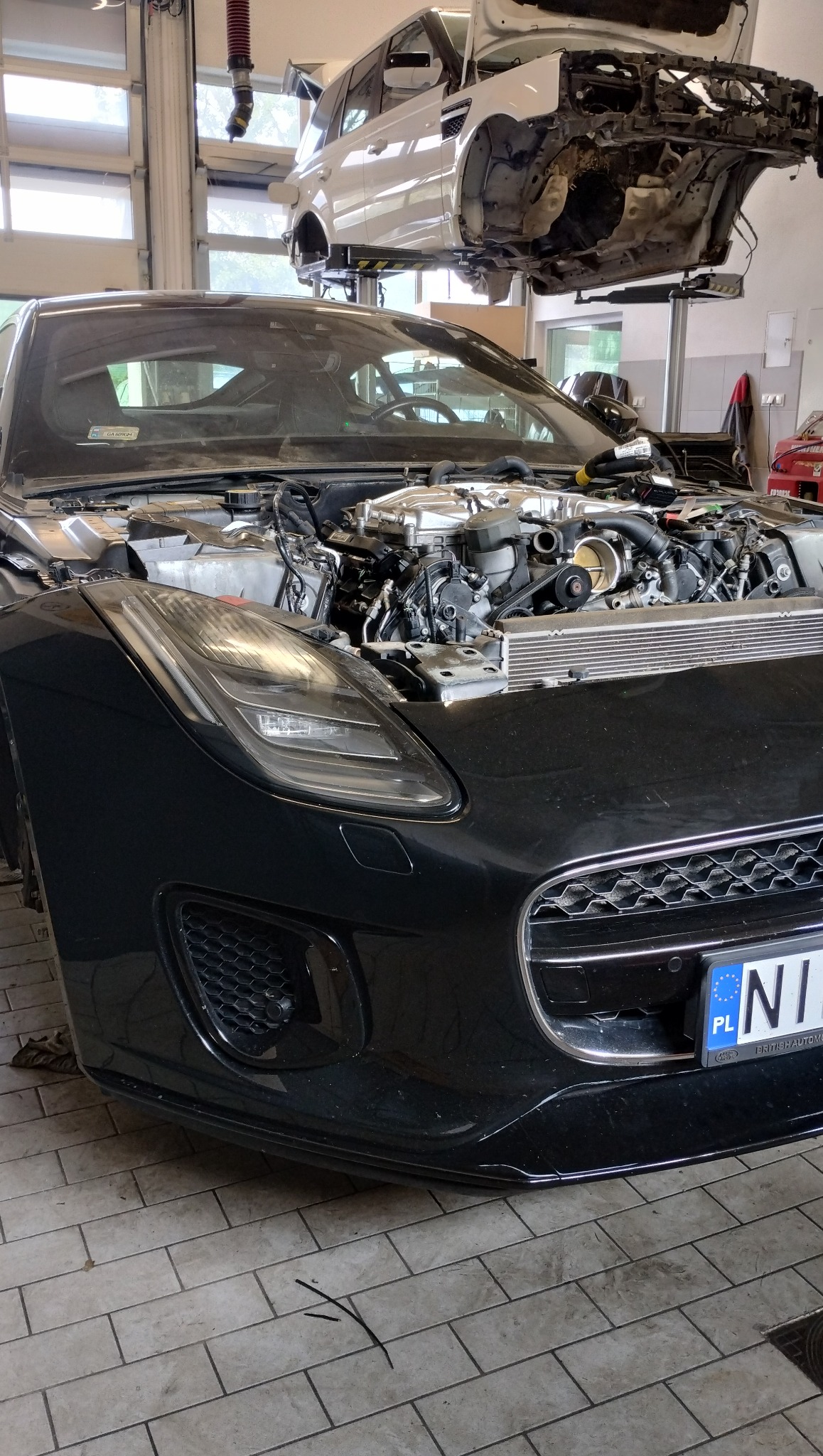 Czarny Jaguar F-Type z otwartą maską w warsztacie, silnik na pierwszym planie, drugi samochód na podnośniku w tle. Widoczne detale silnika i oświetlenie warsztatowe.
