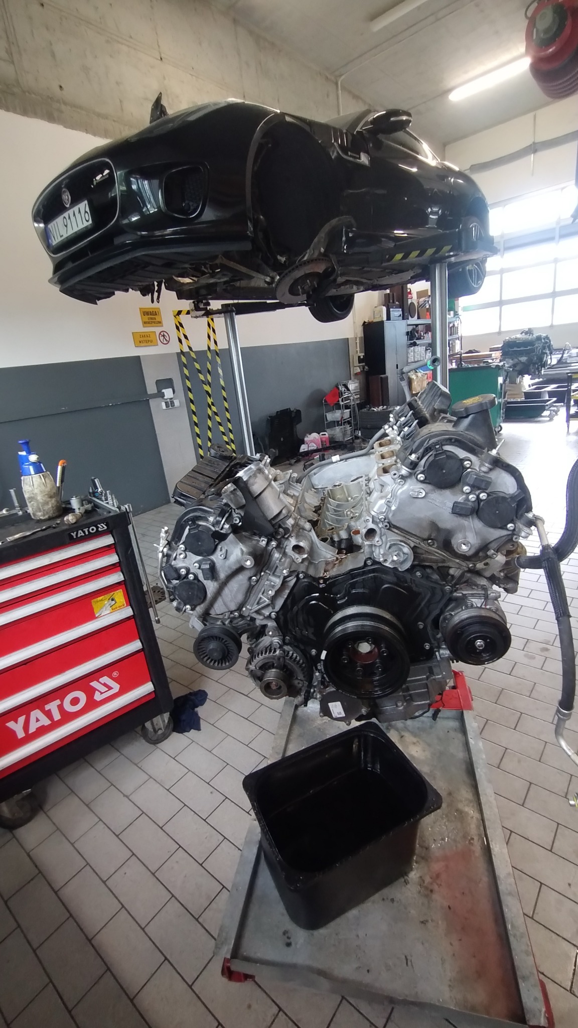 Czarny Jaguar na podnośniku warsztatowym, z wyjętym silnikiem obok. Widoczny wózek narzędziowy Yato i pojemnik na olej. Detal warsztatu samochodowego.