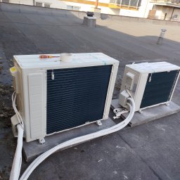 NEVE - Dwie beżowe jednostki zewnętrzne systemu chłodzenia na płaskim dachu, posadowione na betonowych płytach, z białymi rurami instalacyjnymi i narzędziami na większej z nich, w tle budynki.
