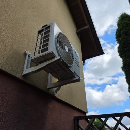 NEVE - Biała jednostka zewnętrzna klimatyzacji z widocznym wentylatorem, zamontowana na beżowo-brązowej elewacji budynku, z kroplą wody spływającą z odpływu, na tle błękitnego nieba i zielonych tui.