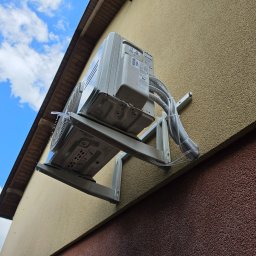NEVE - Biała jednostka zewnętrzna klimatyzacji zamontowana na beżowej ścianie budynku z brązowym cokołem, widoczne rury instalacyjne i krople wody. Ujęcie z dołu na tle błękitnego nieba.