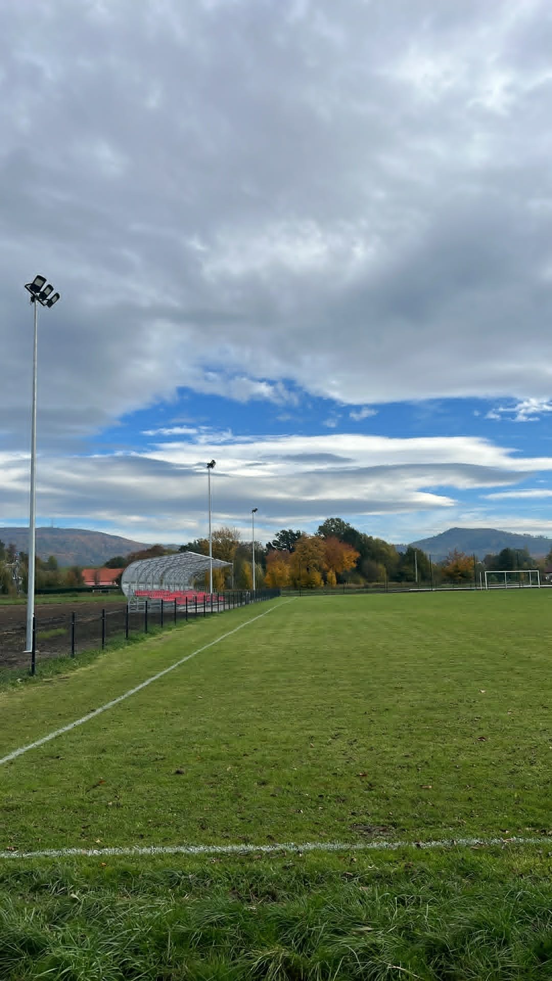 Widok na boisko sportowe z trybunami i ogrodzeniem. W tle wzgórza i pochmurne niebo. Oświetlenie boiska na wysokich masztach.