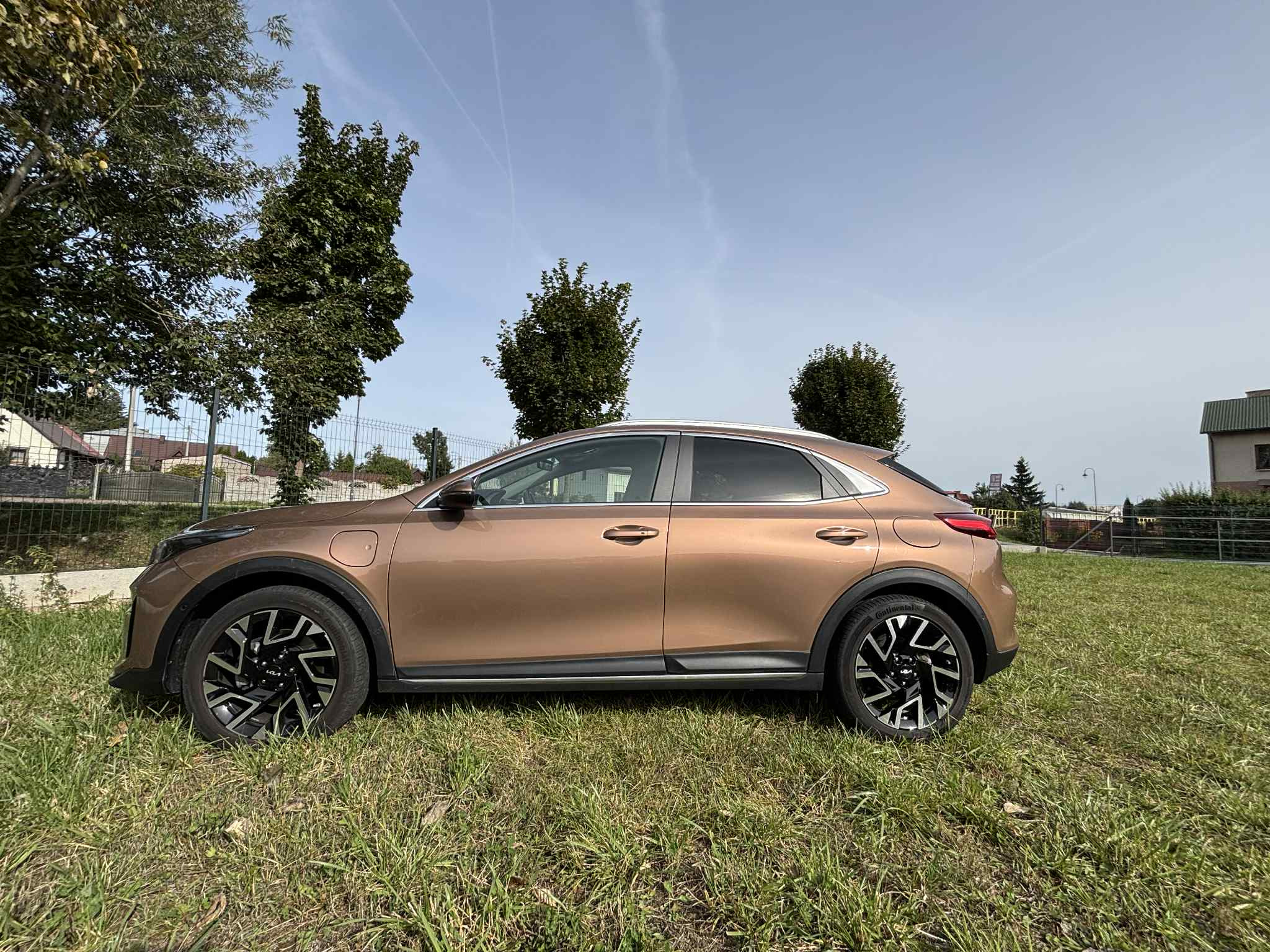 Brązowy crossover Kia XCeed na trawie, widok z boku, felgi aluminiowe z czarnymi akcentami, linia okien w kolorze srebrnym, tło: drzewa i budynki.