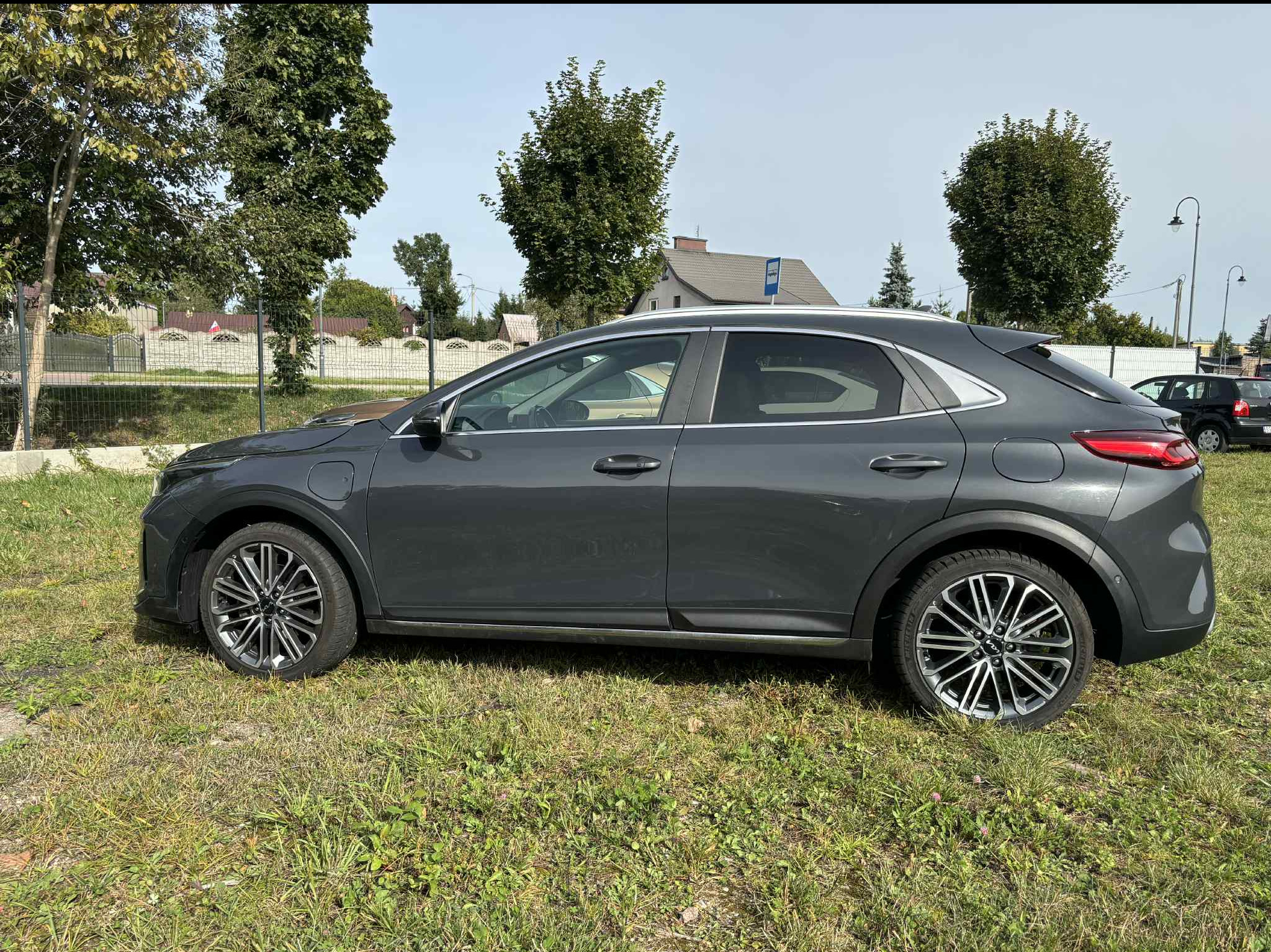 Szary crossover Kia XCeed zaparkowany na trawie, widok z boku, felgi aluminiowe, w tle drzewa i budynek. Auto z wypożyczalni samochodów.