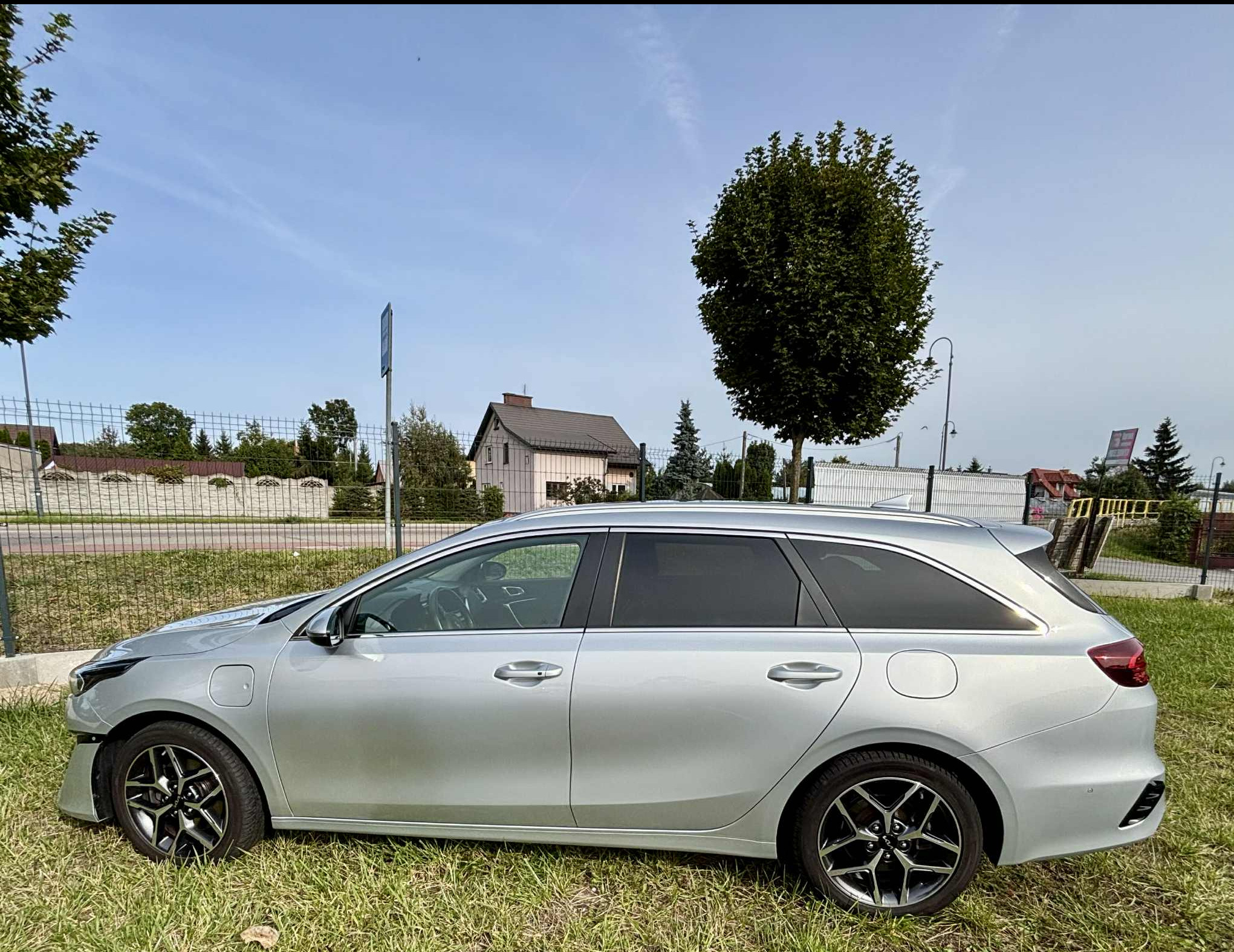 Srebrne kombi Kia Ceed z przyciemnionymi szybami, zaparkowane na trawie w słoneczny dzień, widok z boku. W tle dom i drzewa.