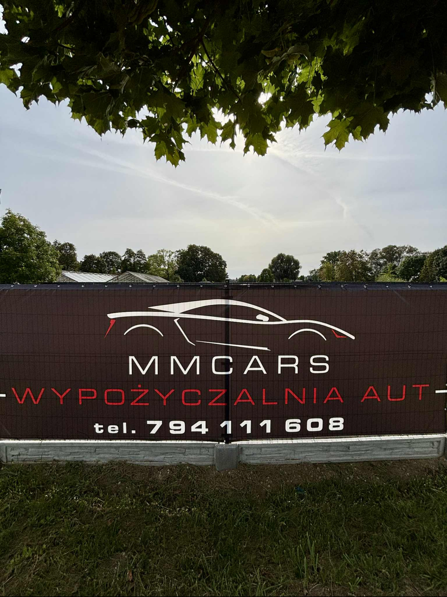 Baner reklamowy wypożyczalni samochodów MM Cars z sylwetką sportowego auta, numerem telefonu i hasłem „Wypożyczalnia Aut” na tle zieleni i błękitnego nieba.