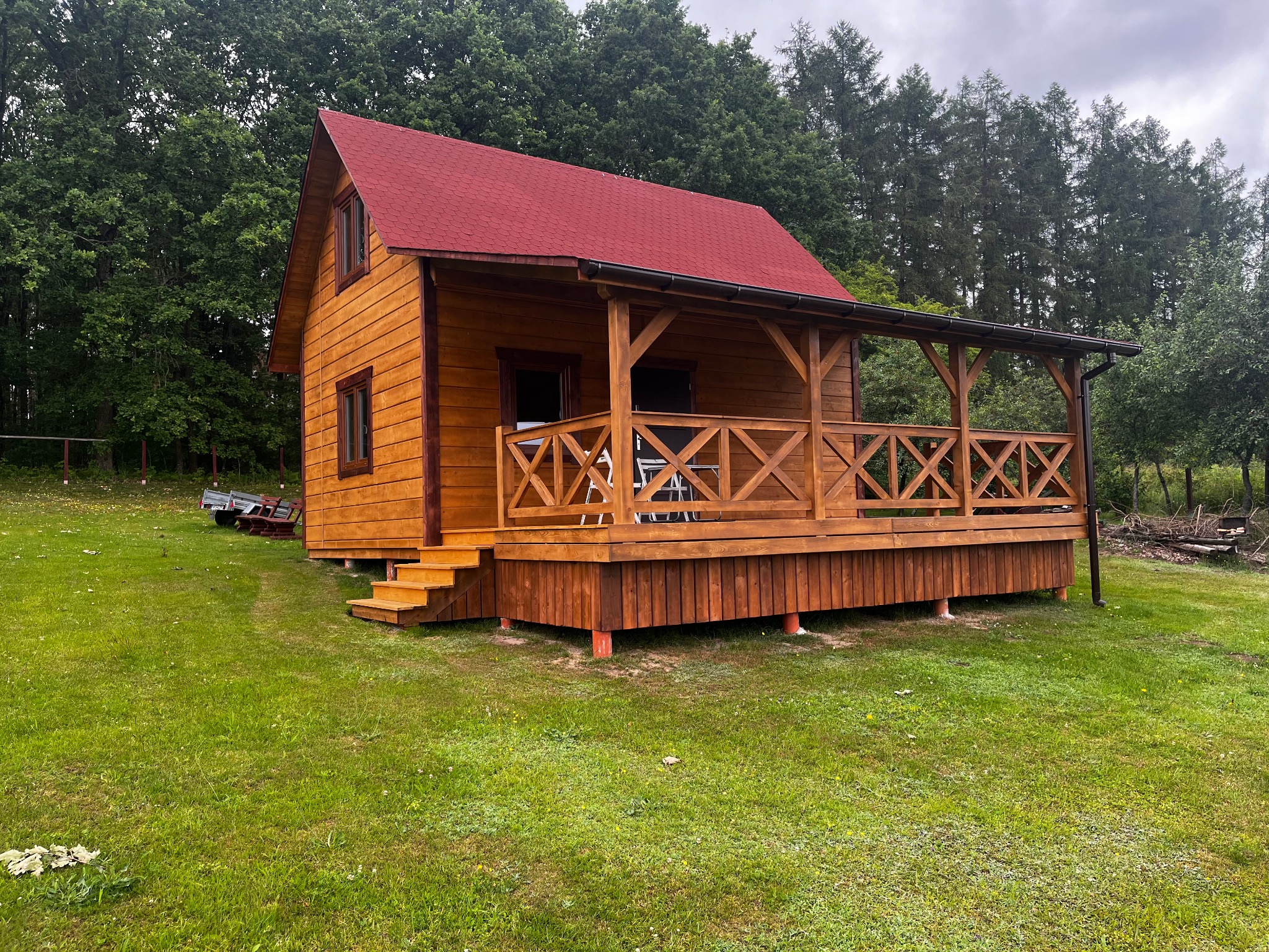 Drewniany domek letniskowy z czerwonym dachem i zadaszonym tarasem, otoczony zielenią. Widoczny fragment przyczepy z tyłu. Całość w naturalnym otoczeniu.