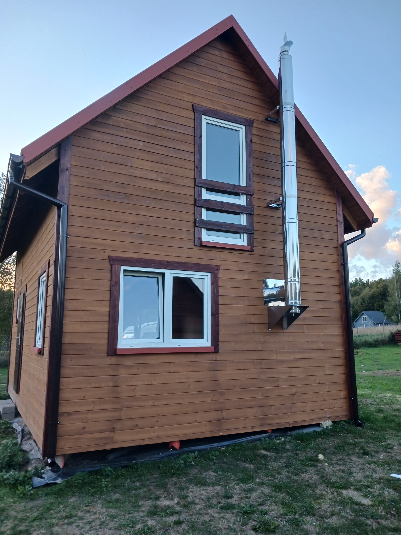 Brązowy dom szkieletowy z kominem ze stali nierdzewnej, widok z boku na tle zieleni i błękitnego nieba. Charakterystyczne okna z białymi ramami.