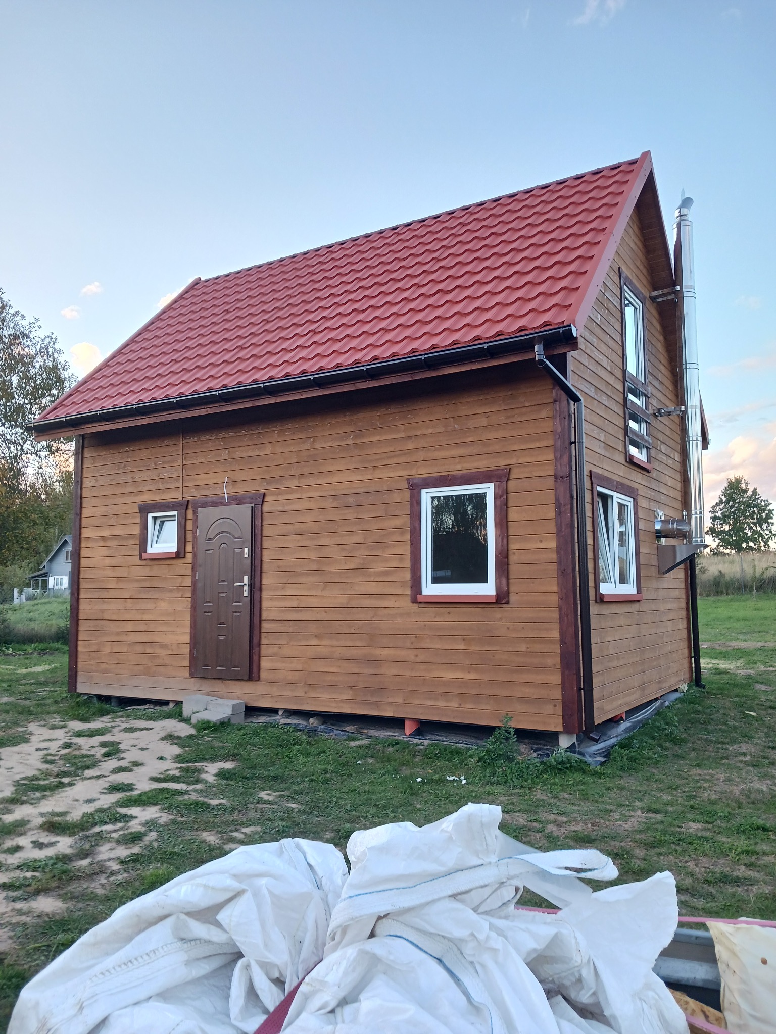 Niewielki, brązowy dom szkieletowy z czerwonym dachem z blachodachówki i metalowym kominem. Widoczne białe okna i brązowe drzwi. W tle zielona trawa i niebieskie niebo.