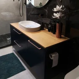 Nowoczesna szafka łazienkowa z drewnianym blatem, umywalką nablatową i czarnymi frontami z miedzianymi uchwytami. Minimalistyczny design, ciemne marmurowe tło.