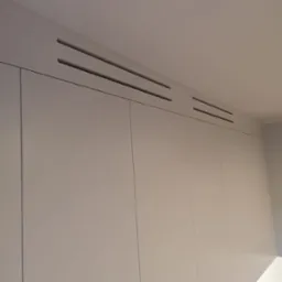 Minimalistyczna szafa wnękowa z gładkimi, jasnymi frontami i subtelnymi, poziomymi szczelinami wentylacyjnymi pod sufitem.