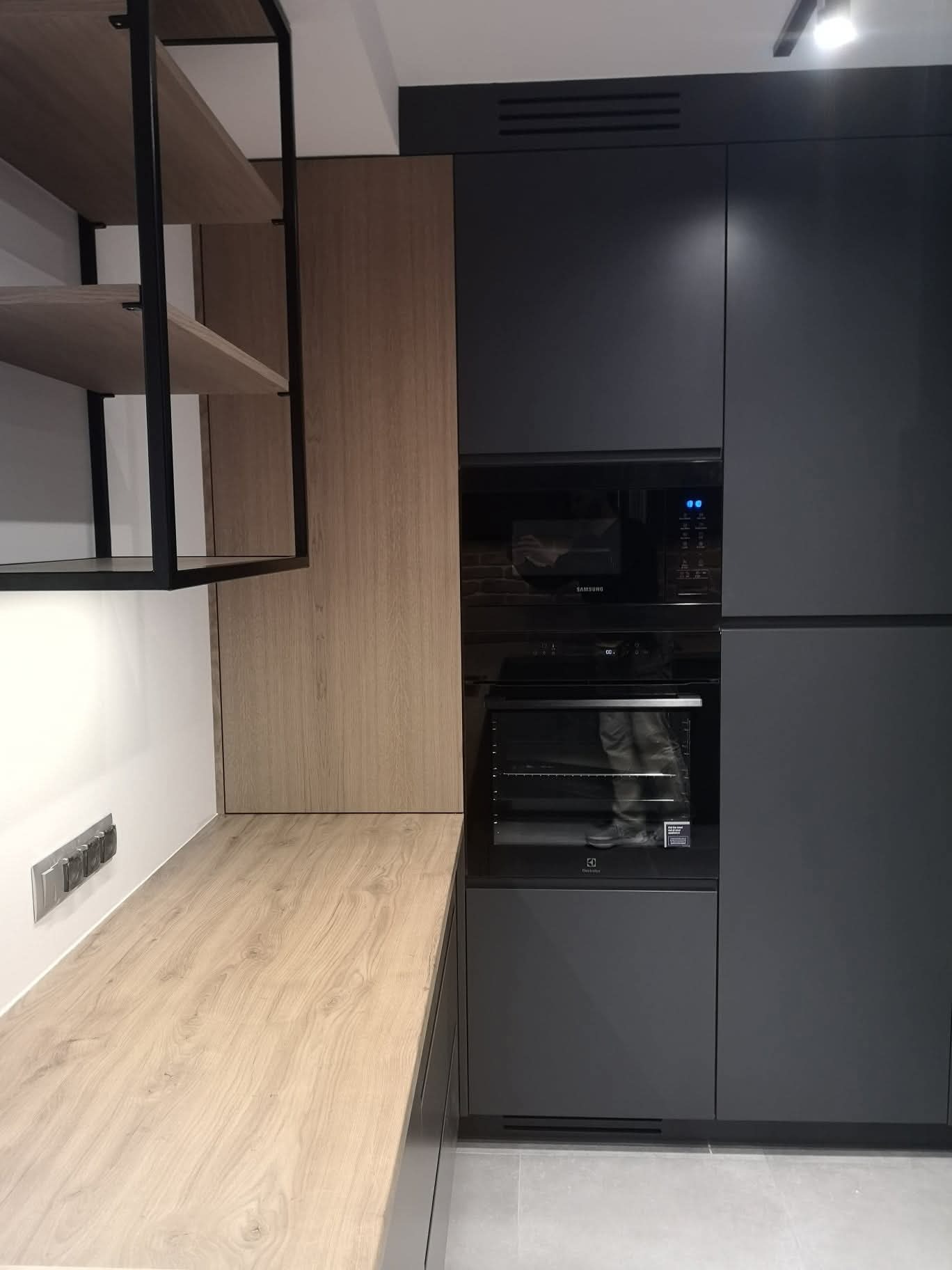 Nowoczesna kuchnia z zabudową w odcieniach szarości i drewna. Widoczne sprzęty AGD w zabudowie oraz półki z czarnymi, metalowymi ramami. Minimalistyczny design.