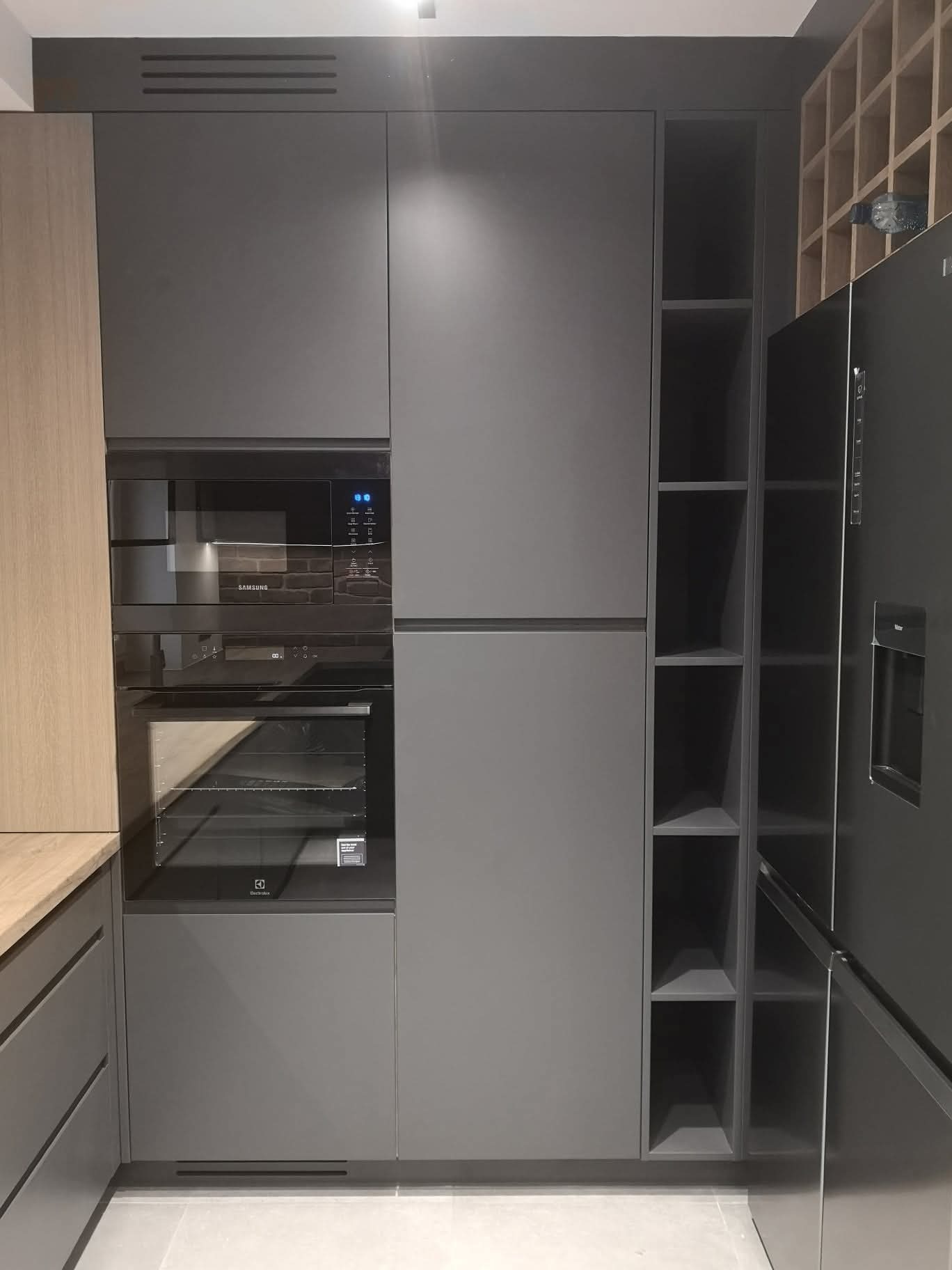 Nowoczesna kuchnia z szarymi frontami, zabudowaną lodówką i piekarnikiem. Obok drewniany regał na wino. Minimalistyczny design, oświetlenie punktowe.