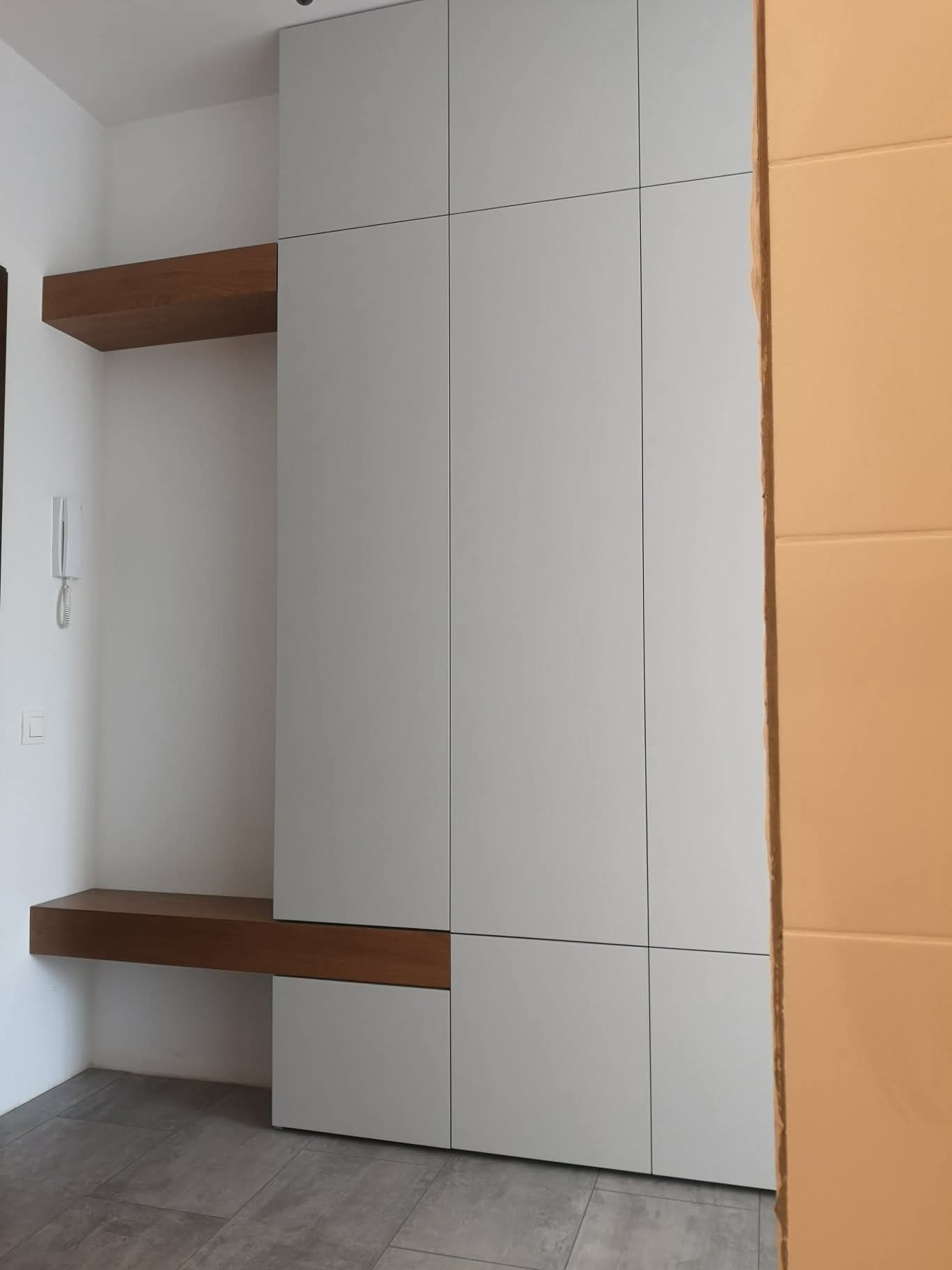 Nowoczesna szafa wnękowa z drewnianymi półkami, minimalistyczny design, szare fronty, widok z boku, szara podłoga, element pomarańczowej ściany w tle.