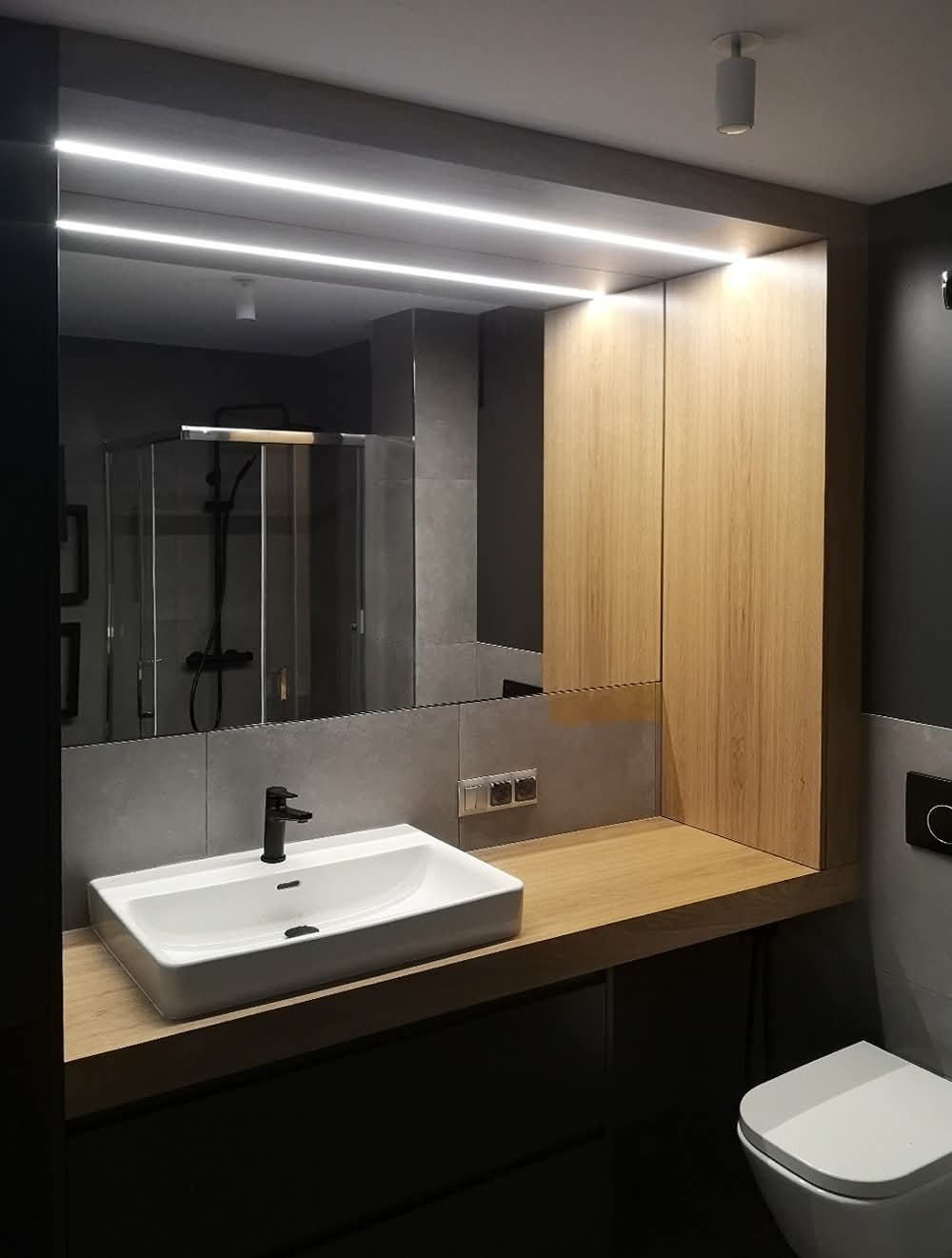 Nowoczesna łazienka z drewnianą szafką i blatem, białą umywalką oraz podświetlanym lustrem. W tle widoczna kabina prysznicowa.