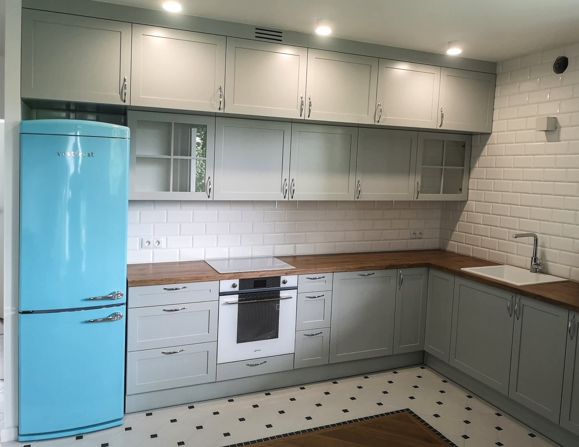 Jasnoszara kuchnia z drewnianym blatem i błękitną lodówką w stylu retro. Białe płytki na ścianie i wzorzysta podłoga dodają uroku.