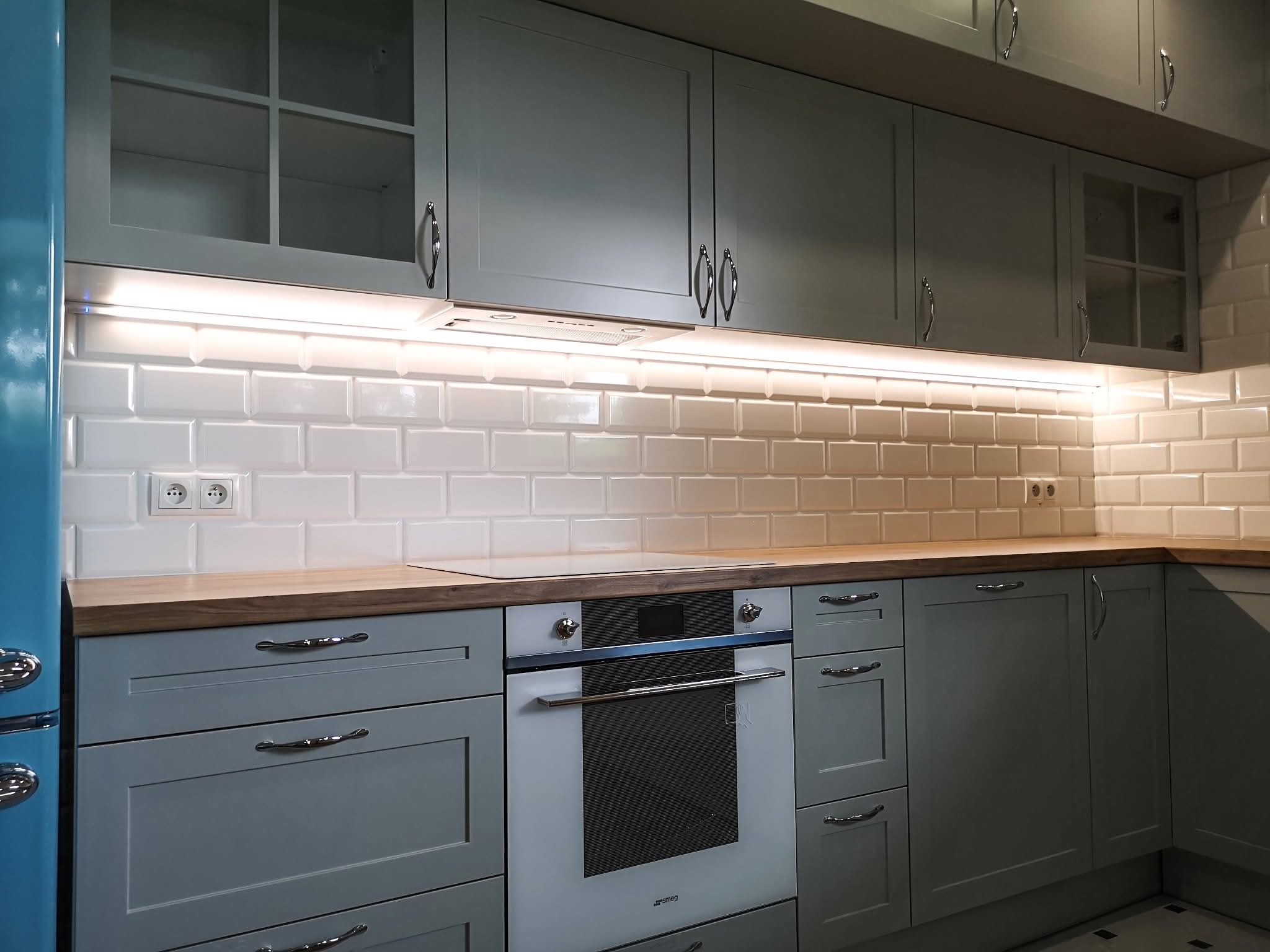 Szare szafki kuchenne z chromowanymi uchwytami i drewnianym blatem. Nad blatem białe kafelki i oświetlenie LED. Widoczny fragment błękitnej lodówki.