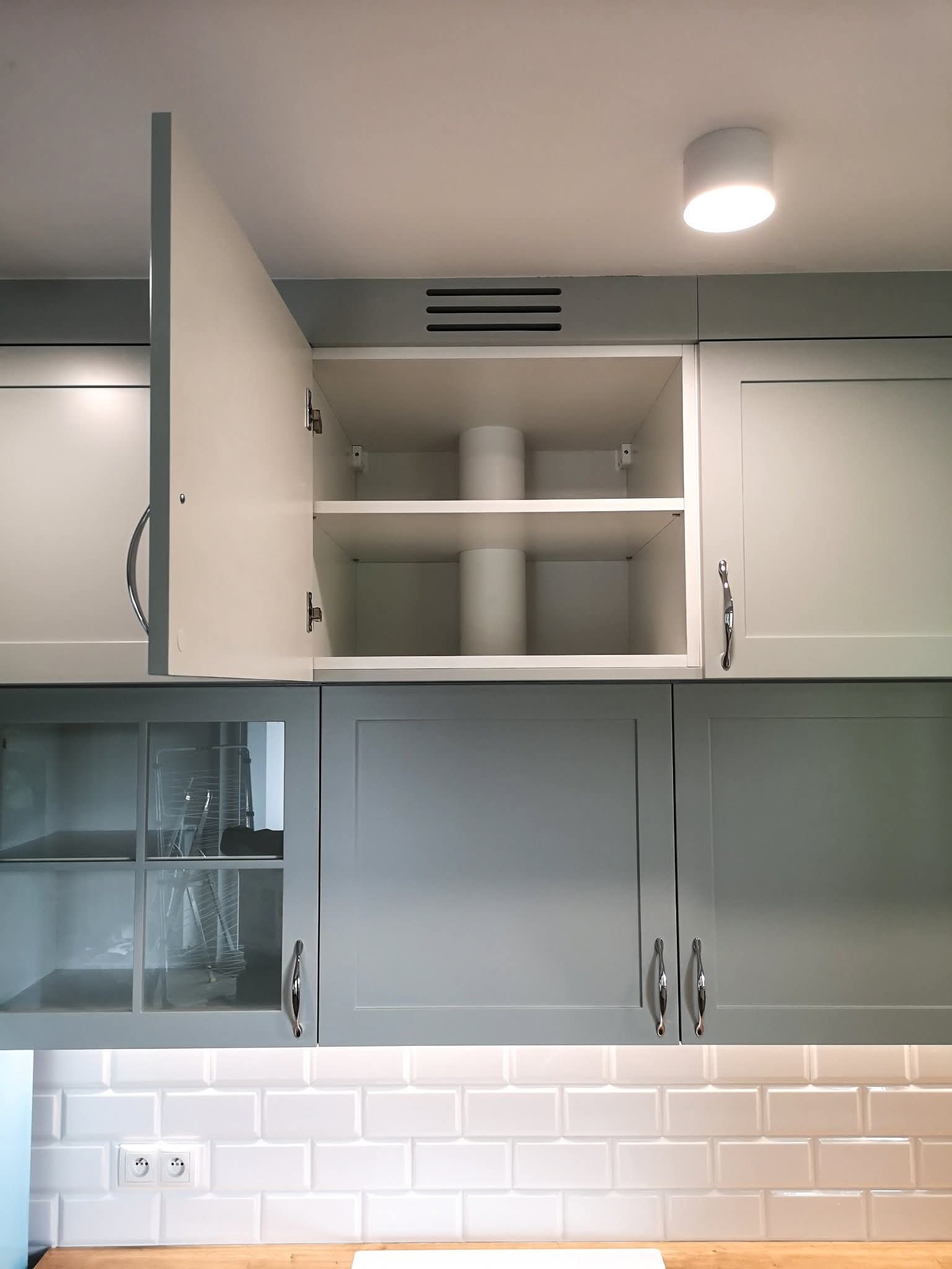 Szare szafki kuchenne z białymi frontami, jedna szafka otwarta, widoczne półki z cylindrycznymi elementami. Nad blatem białe płytki.