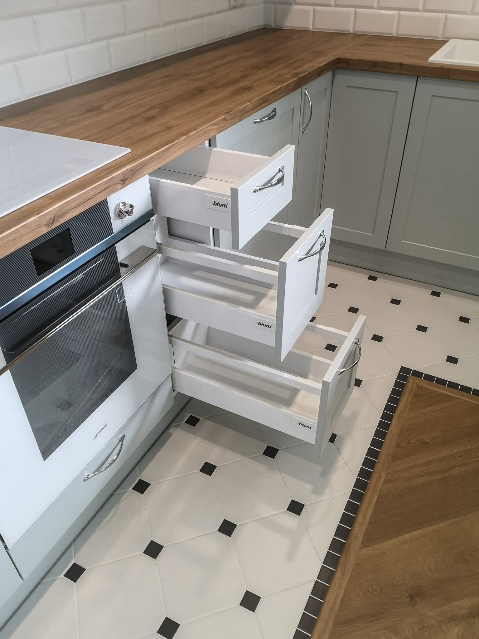 Nowoczesna kuchnia z szarymi szafkami i drewnianym blatem. Widoczne wysuwane szuflady Blum obok piekarnika Smeg. Podłoga w biało-czarną szachownicę.