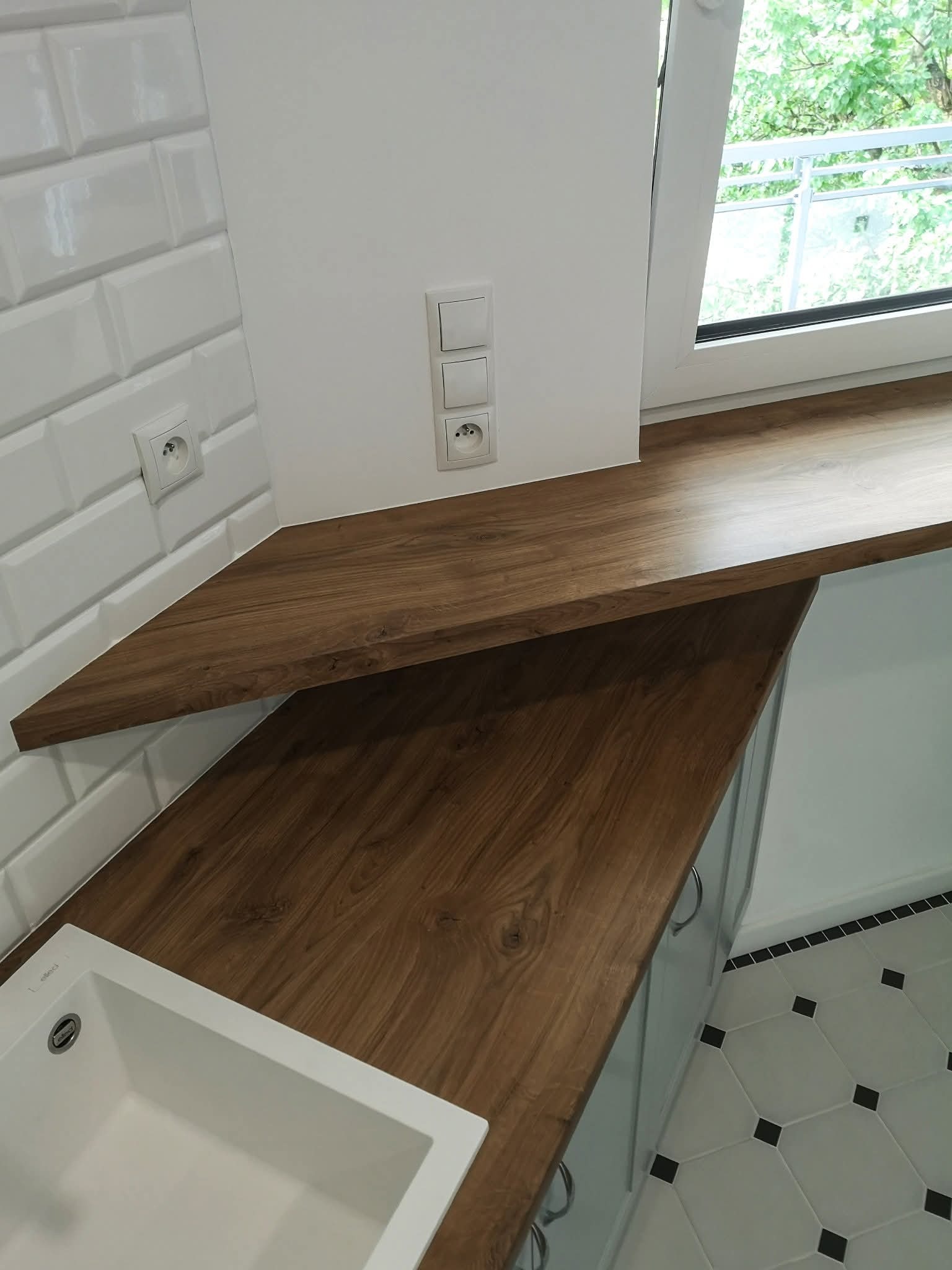 Nowoczesna kuchnia: drewniany blat narożny z wbudowanym zlewem, białe płytki metro i gniazdka elektryczne. Widoczny fragment szafki i wzorzysta podłoga.