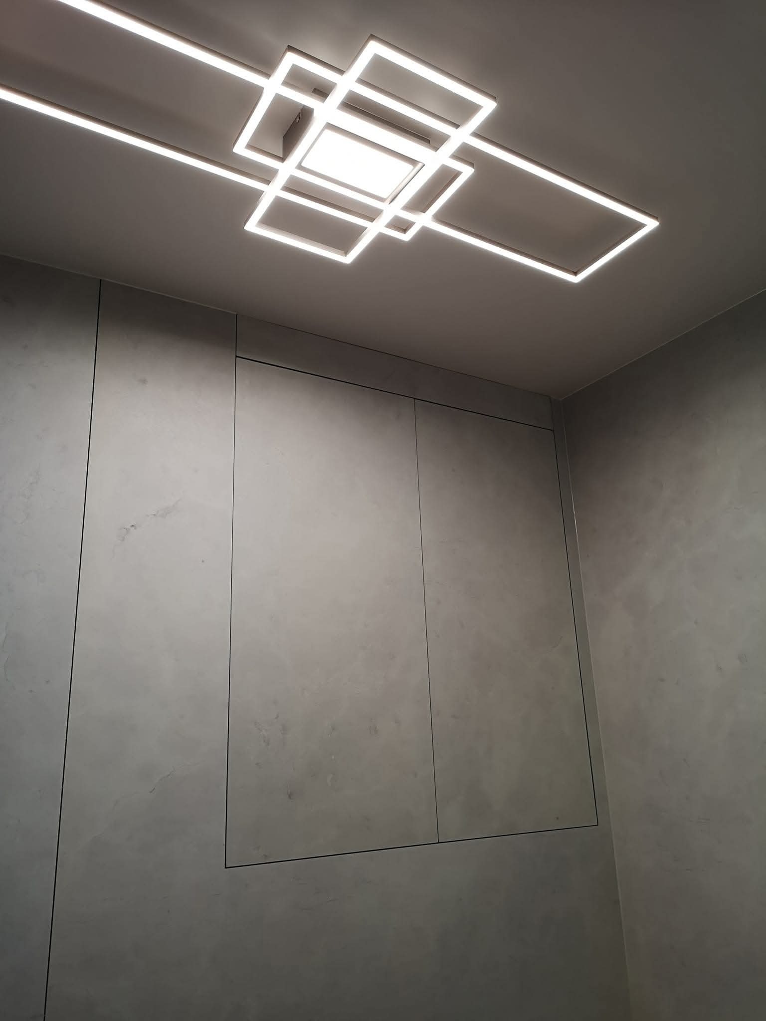 Minimalistyczna szafa wnękowa z szarymi frontami, wkomponowana w ścianę o fakturze betonu, oświetlona nowoczesną lampą sufitową.