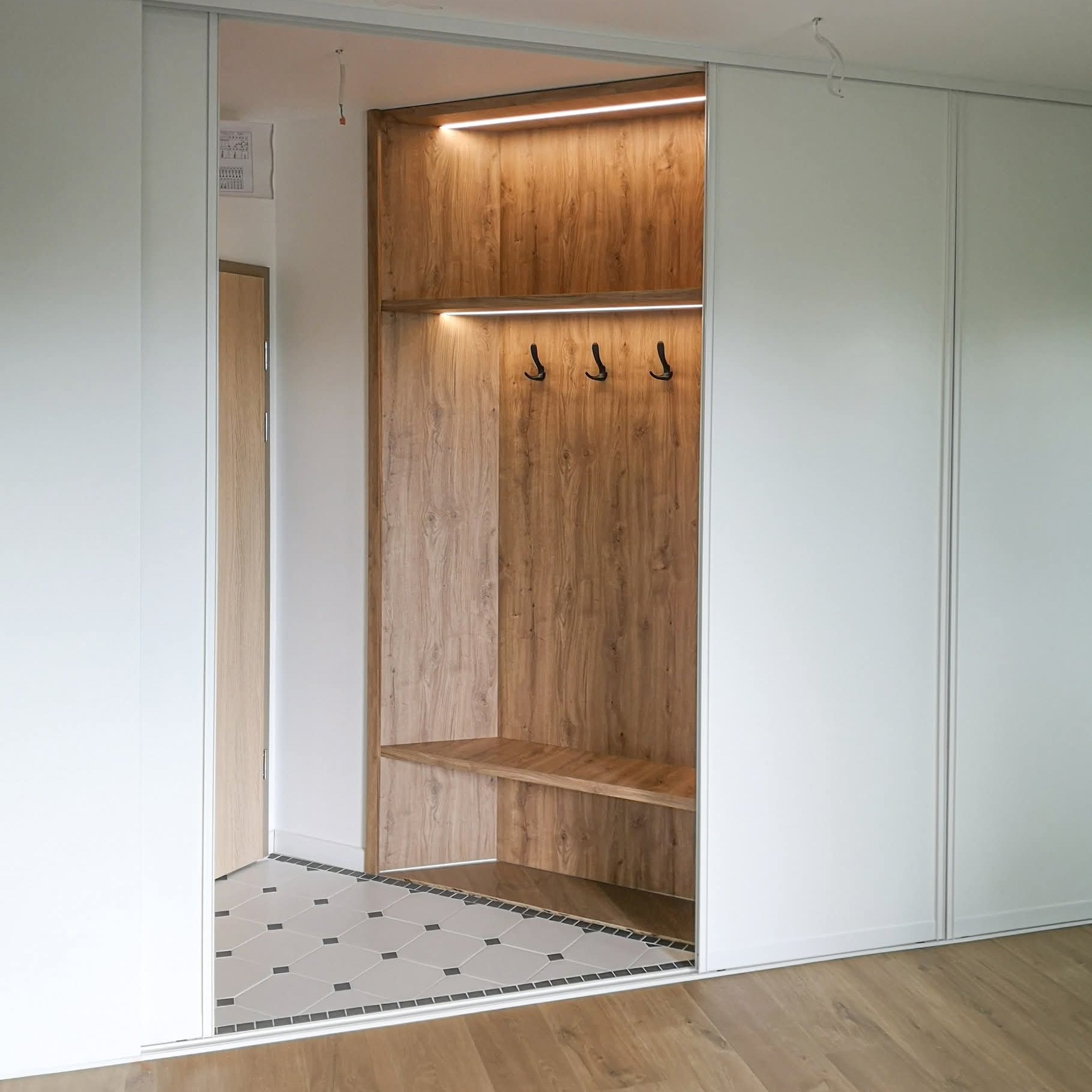 Wnęka z drewnianym wykończeniem, oświetleniem LED i haczykami na ubrania, wkomponowana w białą szafę przesuwną. Na podłodze wzorzyste płytki.