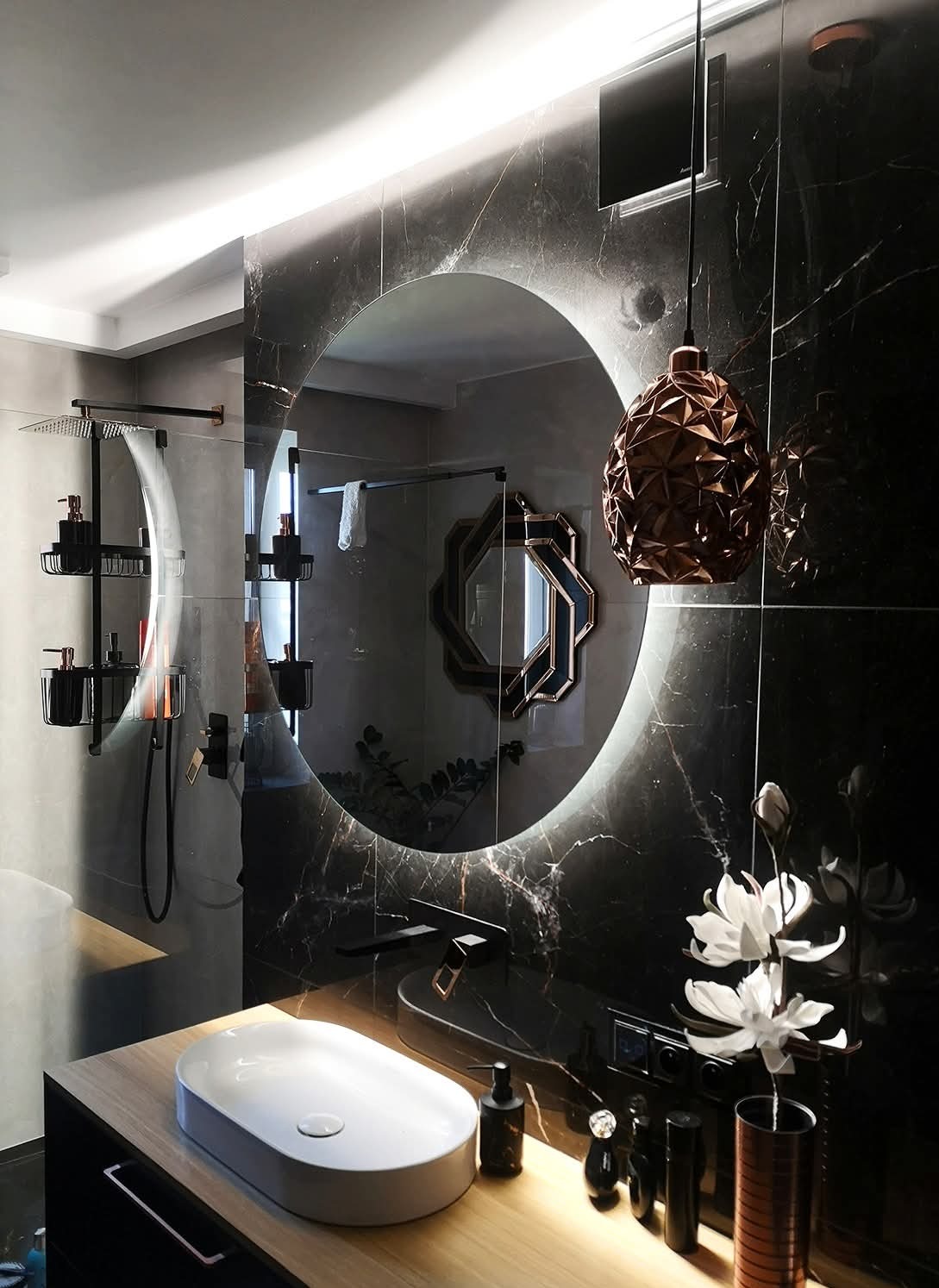 Elegancka łazienka w Raszynie z okrągłym lustrem LED, czarnymi płytkami marmurowymi i drewnianą szafką pod umywalkę. Nowoczesny design z kwiatami i lampą.